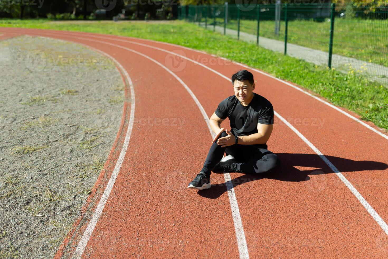 Sports injury. A young Asian man is sitting at the stadium on a treadmill, holding his leg and knee. He writhes in pain. Needs medical help. photo