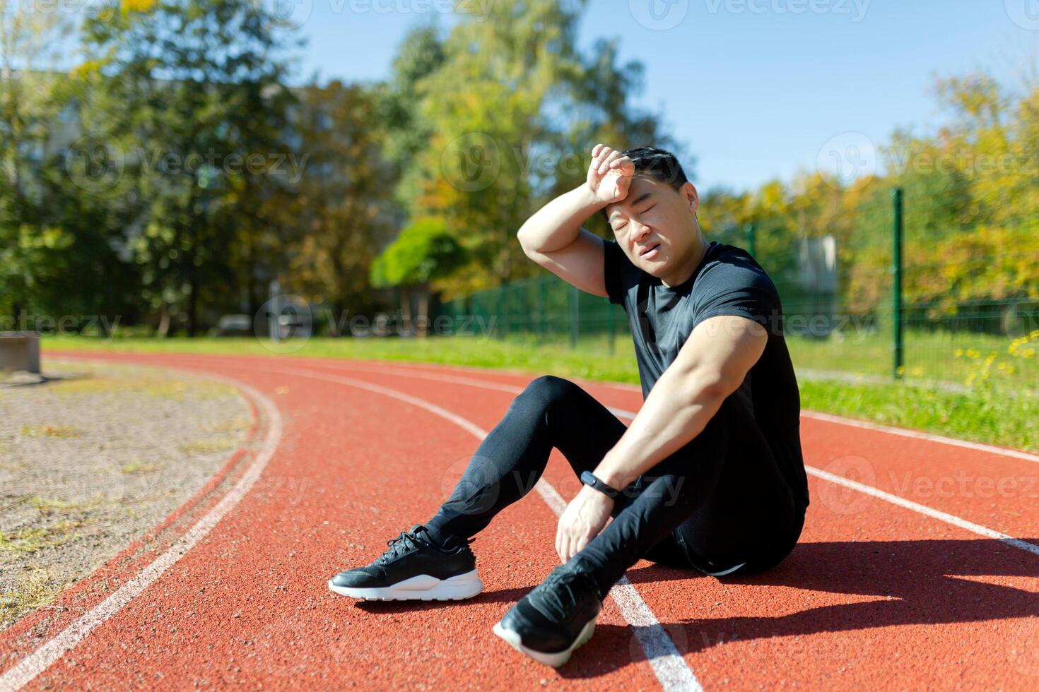 Tired young asian sportsman and runner sitting in stadium and wiping sweat from forehead with hand. photo