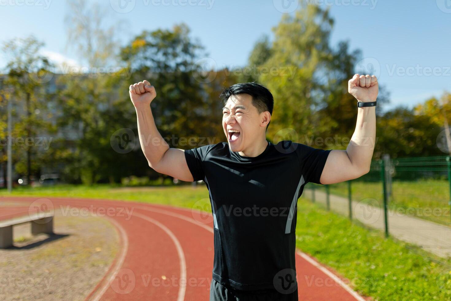 Asian young male runner and sportsman standing in stadium, raising hands up, rejoicing and celebrating victory at finish line, satisfied with result. photo