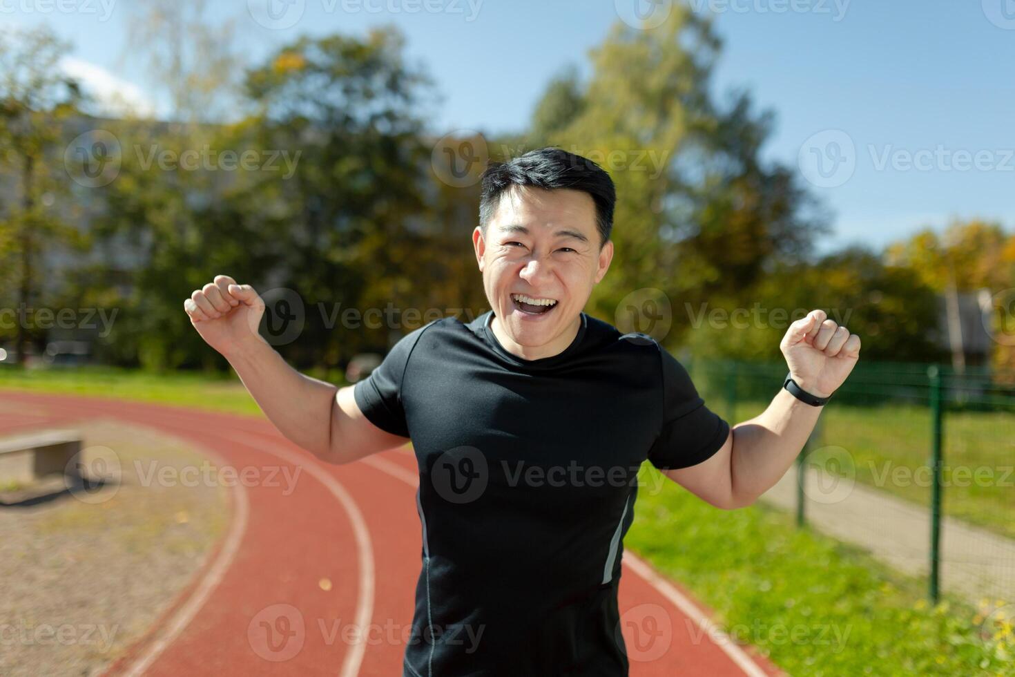 A young Asian man, an athlete, a runner celebrates a victory in a race, in a marathon. The happy man is standing in the stadium on the running track, raised his hands up, rejoices in the camera. photo