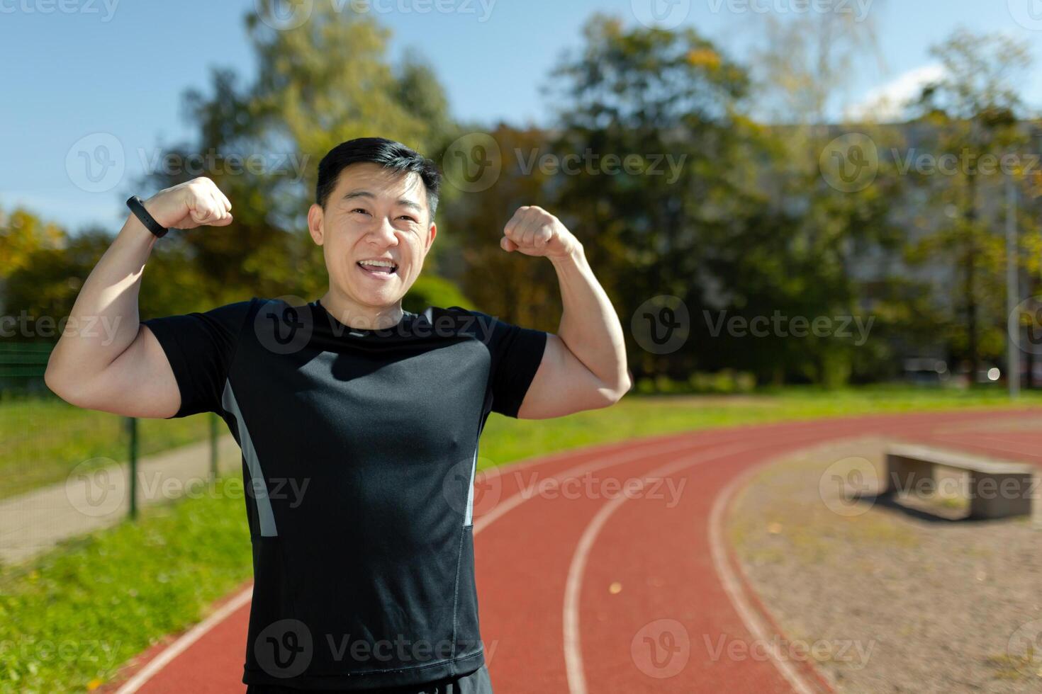 Portrait of an Asian male sportsman standing in a stadium and showing the camera a power gesture with his hands, showing off his toned muscles and body. photo