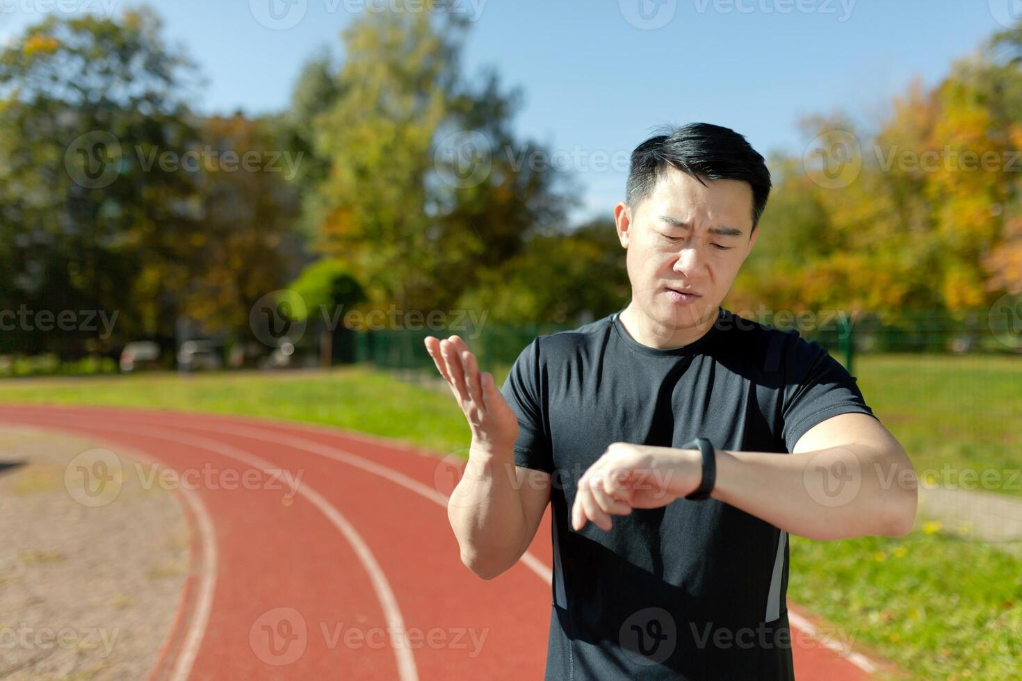 Portrait of a young Asian male athlete, a runner standing on a treadmill in a stadium and checking the time on a smart watch. Dissatisfied with the result, he spreads his hands in frustration. photo