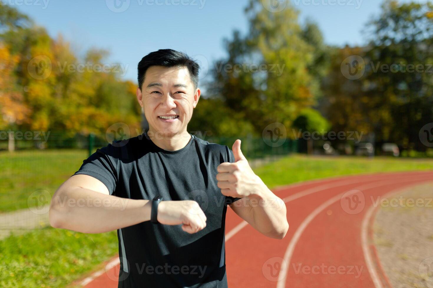 A young Asian man, an athlete standing on a running track in a stadium, checking at the time on a smart watch, pointing a super finger. He smiles, satisfied with the result of the race, marathon. photo