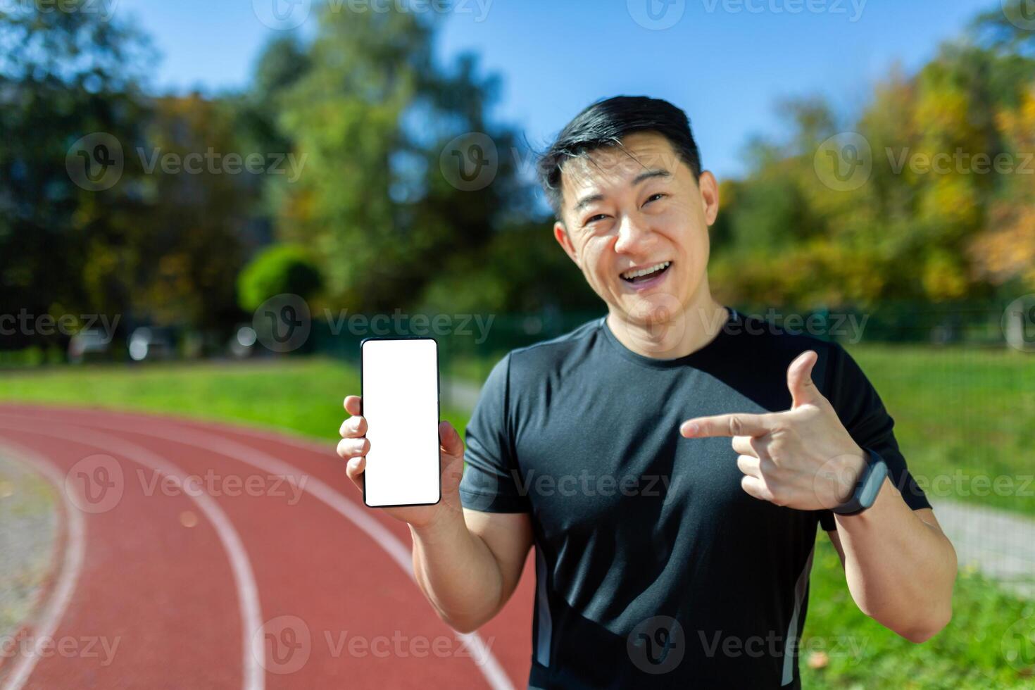 Successful and satisfied Asian athlete on training smiling and looking at camera man showing smartphone screen to camera with blank space on white background, athlete training on sunny day at stadium photo