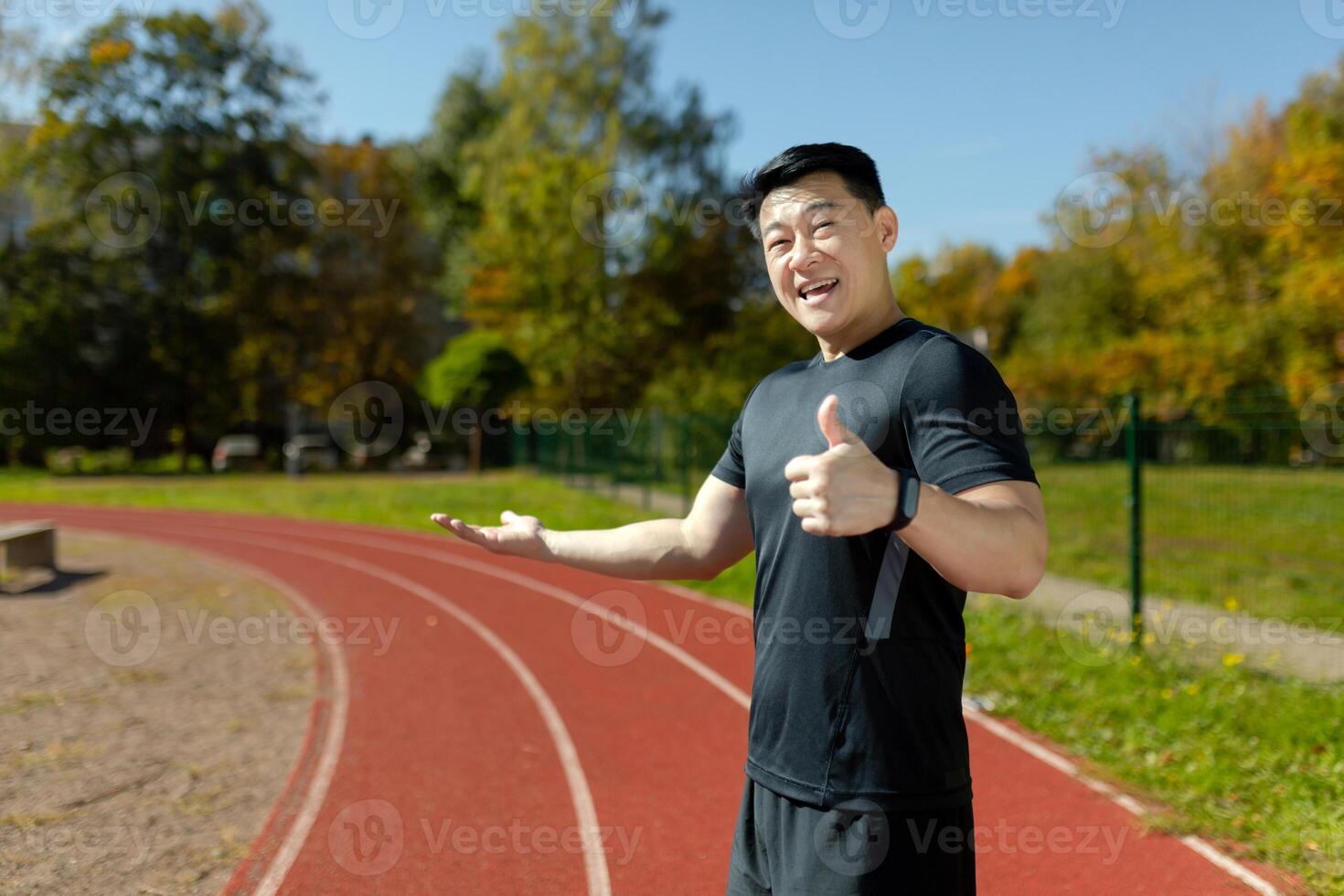 A young Asian male runner is standing in sportswear, at the stadium and is pointing to the running track, with one hand and the super sign with the other hand. photo