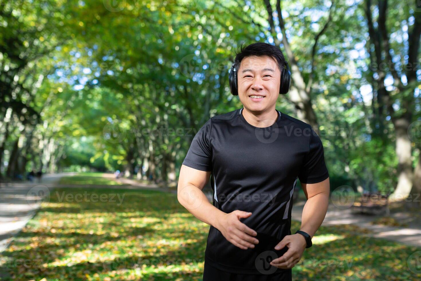 Portrait of a young Asian male athlete running in the park, doing a morning jog in headphones, listening to music. audiobook, podcast. He looks at the camera, smiles. photo