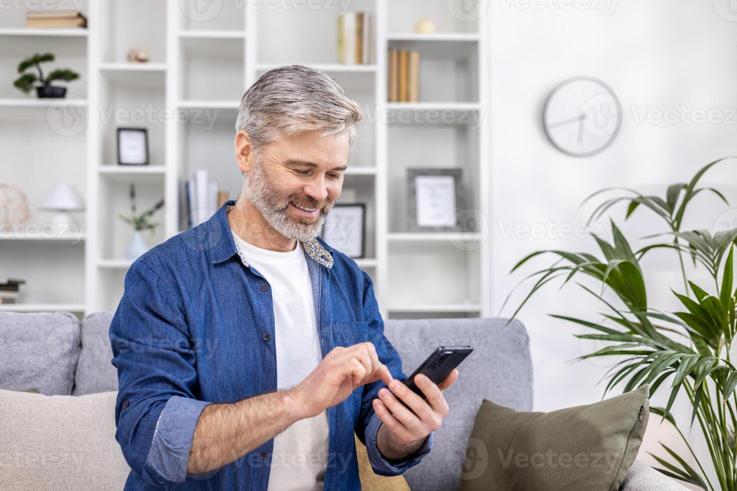 Mature adult man close-up at home smiling and using phone in living room sitting on sofa, person typing message and browsing web page. photo