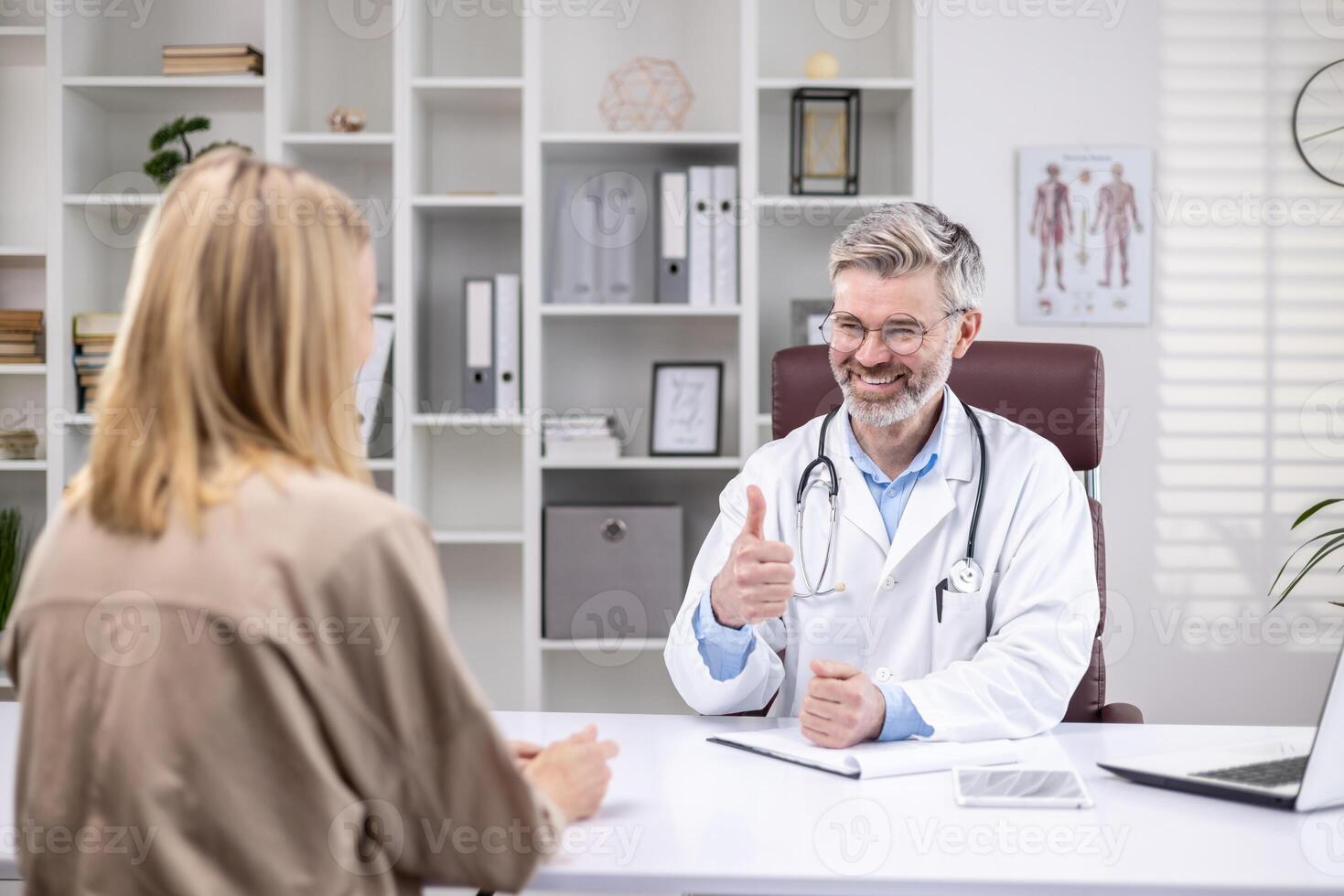 Back view, experienced mature doctor consulting smiling female patient, gray-haired man working inside clinic office, using laptop. photo