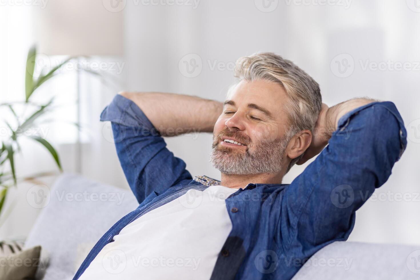 Close-up photo. Senior gray-haired man sitting relaxed on sofa at home and resting, napping. Throwing his hands behind his head, he smiles. photo