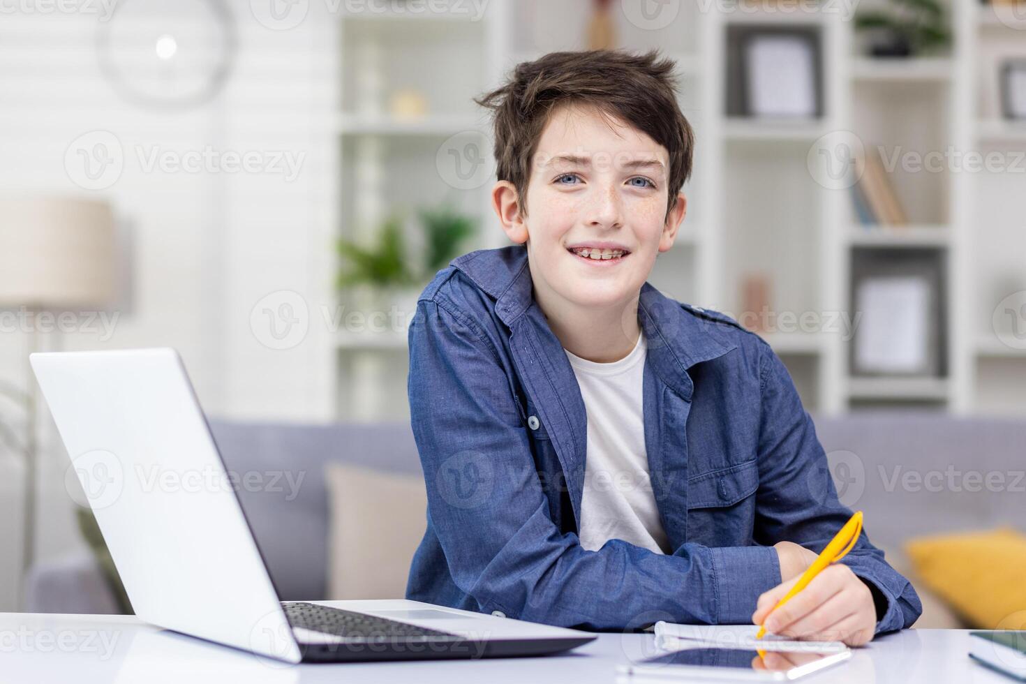 Smiling student boy sitting at a table in a bright room, studying remotely, doing homework, writing in a notebook, smiling, looking at the camera. photo