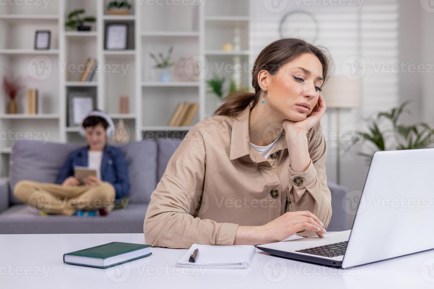 Tired businesswoman working on laptop at home remotely, holding head with hand, tapping on keyboard, programming, mother at work with child in background. photo