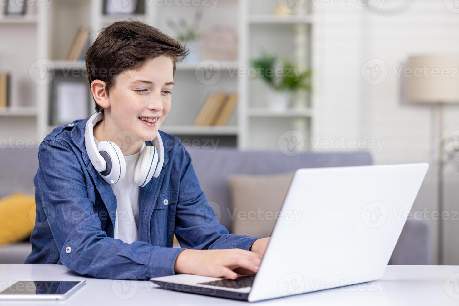Happy teenage boy with headphones around his neck, sitting at a laptop in a bright room, tapping on a keyboard, playing games, monitoring social networks, doing homework. photo