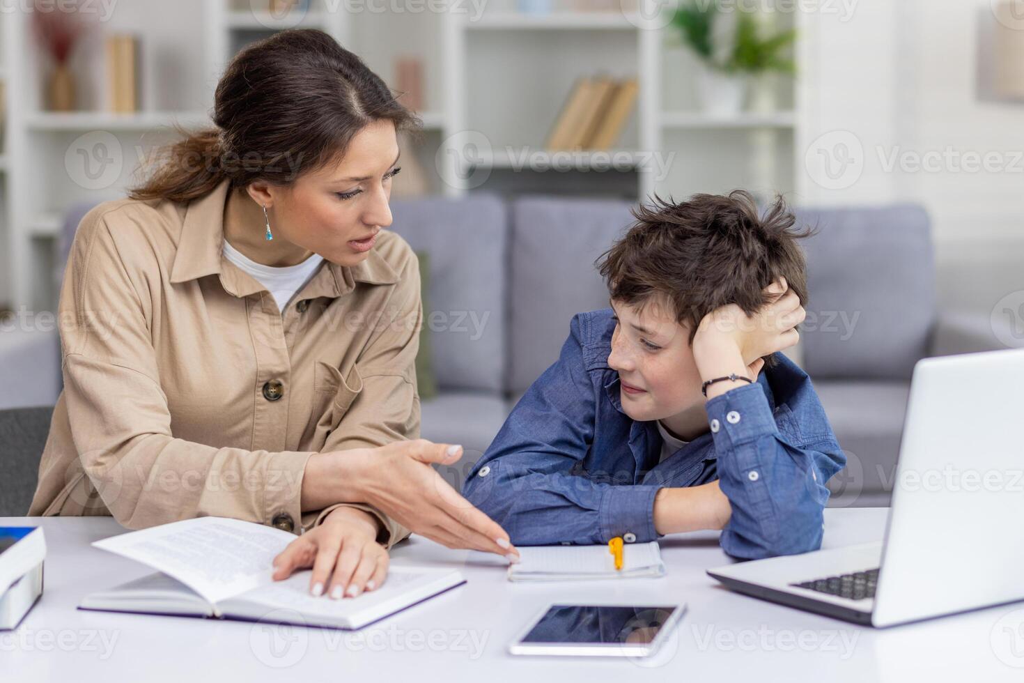 A female tutor works with a boy student in a bright living room at home, schoolboy is tired and dissatisfied with his studies, leans on his hand, does not listen his mother, who helps with homework. photo