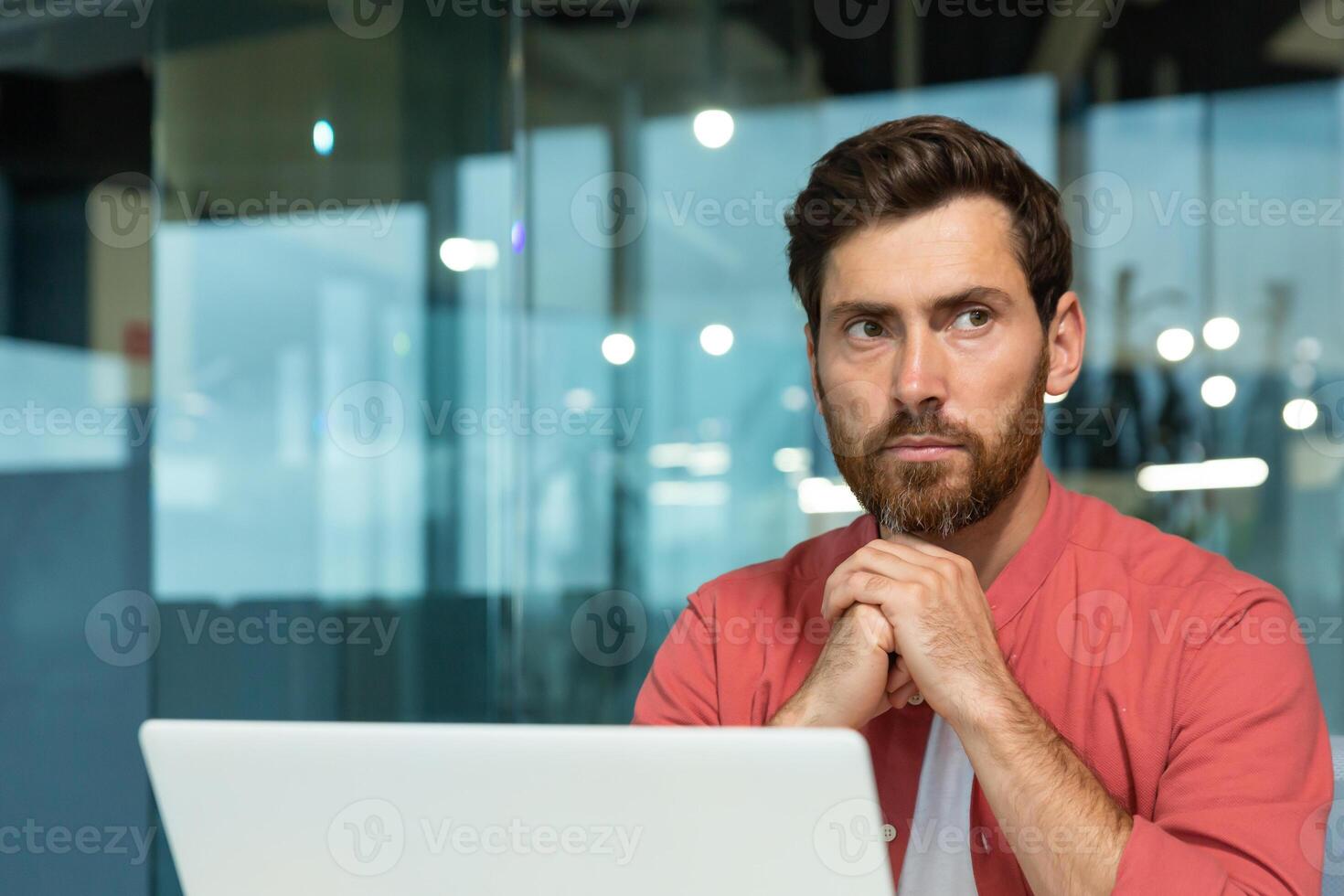 Close-up photo. A young male programmer, designer, freelancer in a red shirt who sits and works in the office at a laptop. He folded his hands together, looks thoughtfully and seriously to the side. photo