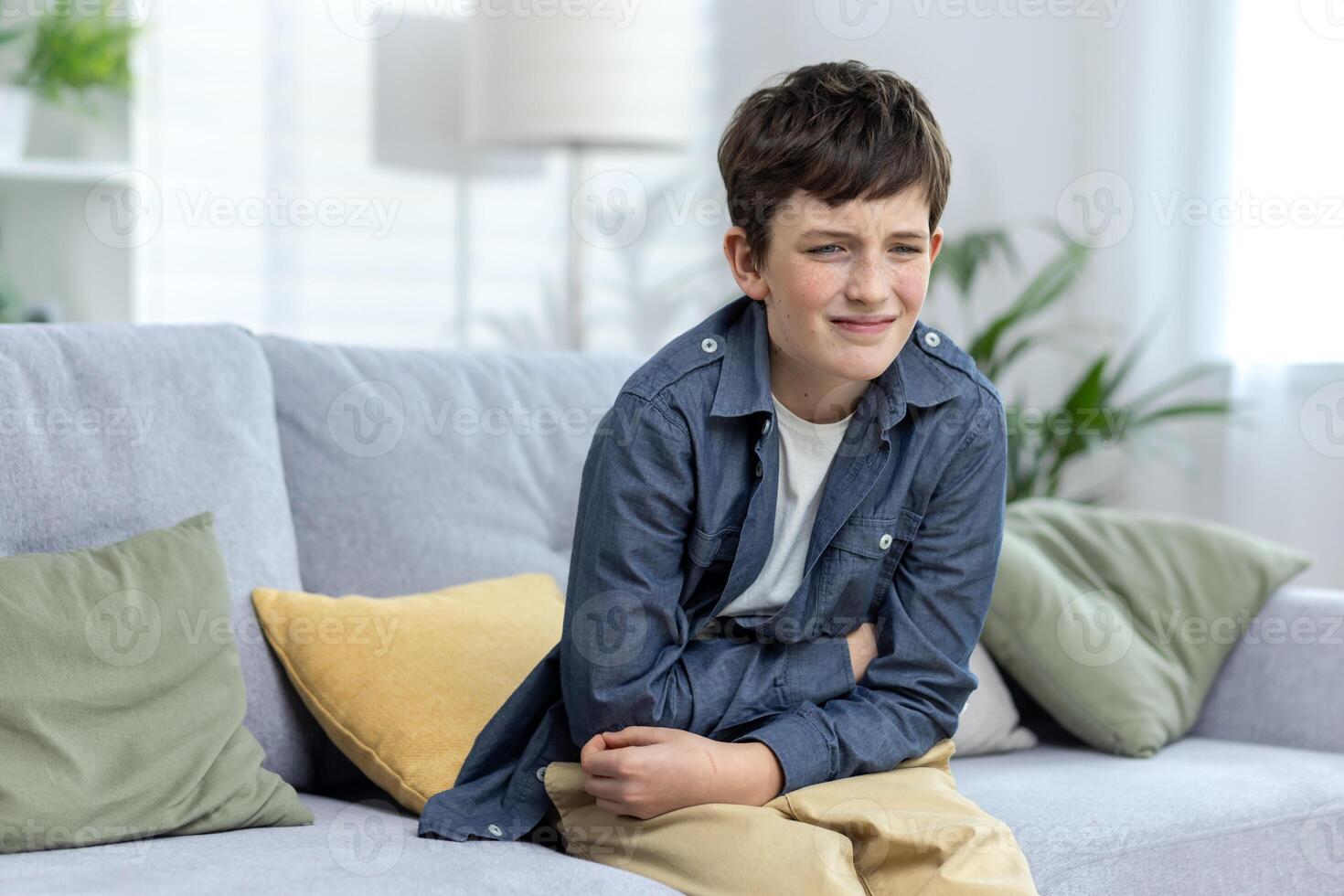 A child, a boy, a teenager sits alone at home on the couch and holds his stomach with his hands. Grimaced, bent, suffering from pain. Poisoned, injured, intestinal infection. appendicitis. photo