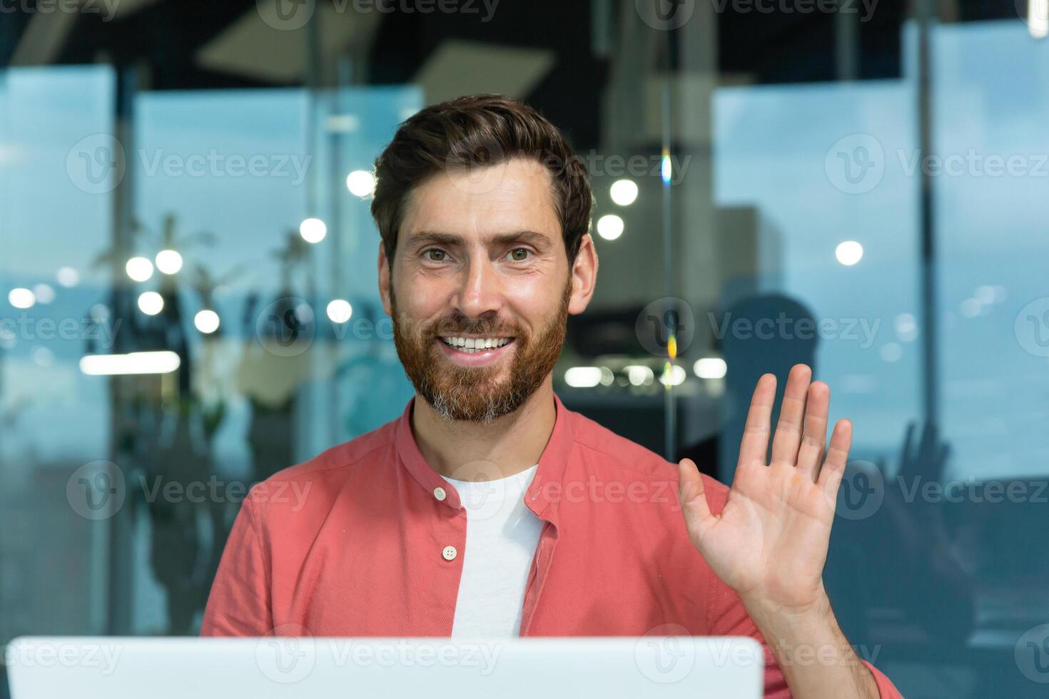 Portrait of a young male programmer in a red shirt working in the office at a laptop. He looks at the camera with a smile, waves his hand. Close-up photo. photo