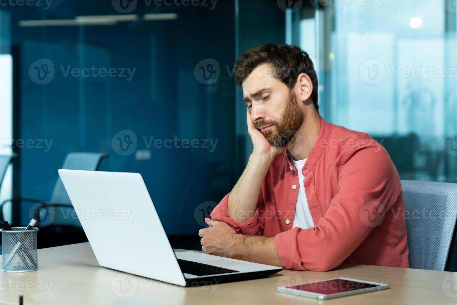 Overtime, deadline. Tired young male programmer, freelancer sits in the office and tries to work at the laptop. Bored, he sits at the table and rests his head on his hand. photo