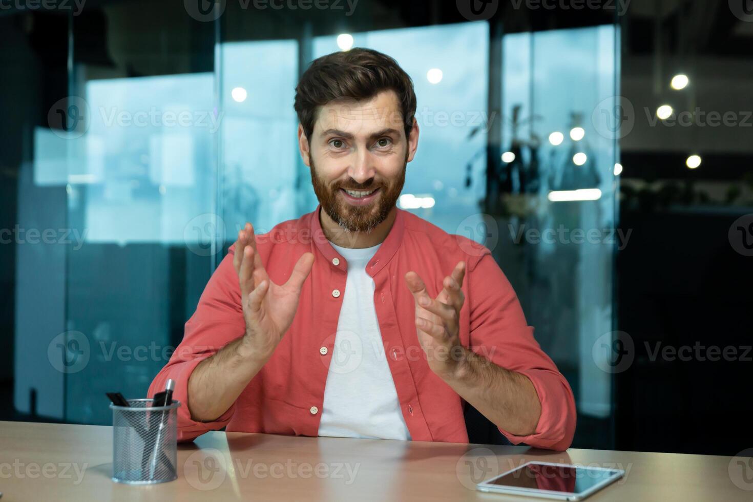 A young male teacher teaches online, conducts lectures, lessons. Sitting in the office at the table in front of the camera. Talks on a call, explains, smiles. photo