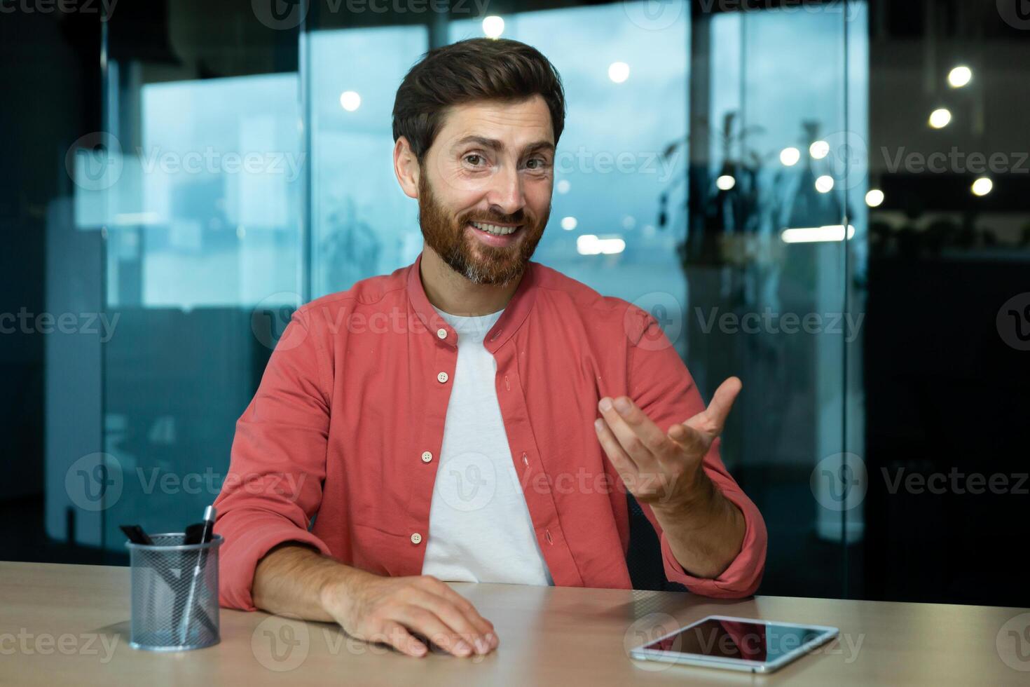 Portrait of a smiling young man sitting at the desk in the office in front of the camera and talking via call, conducting business training, online conference, webinar, business meeting. photo