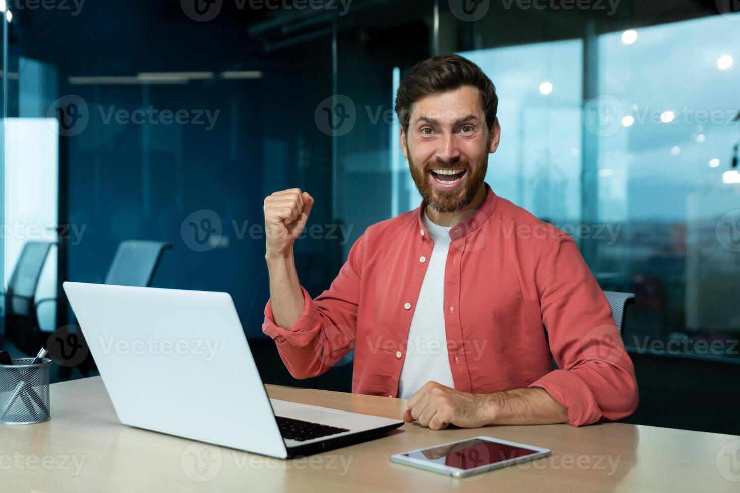 Happy young student sitting in office, campus in front of .aptop, showing victory success gesture with hand, celebrating, looking at camera, smiling. photo