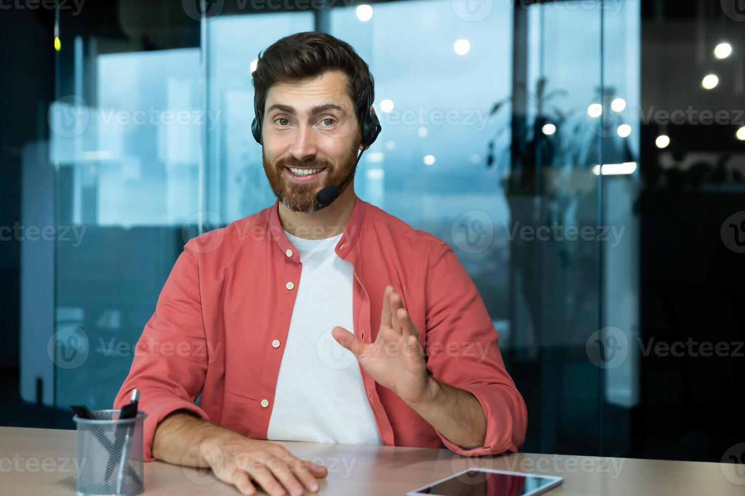 A young man in a red shirt in a headset sits in the office at the table and conducts business training on camera. Explains, advises, smiles. photo