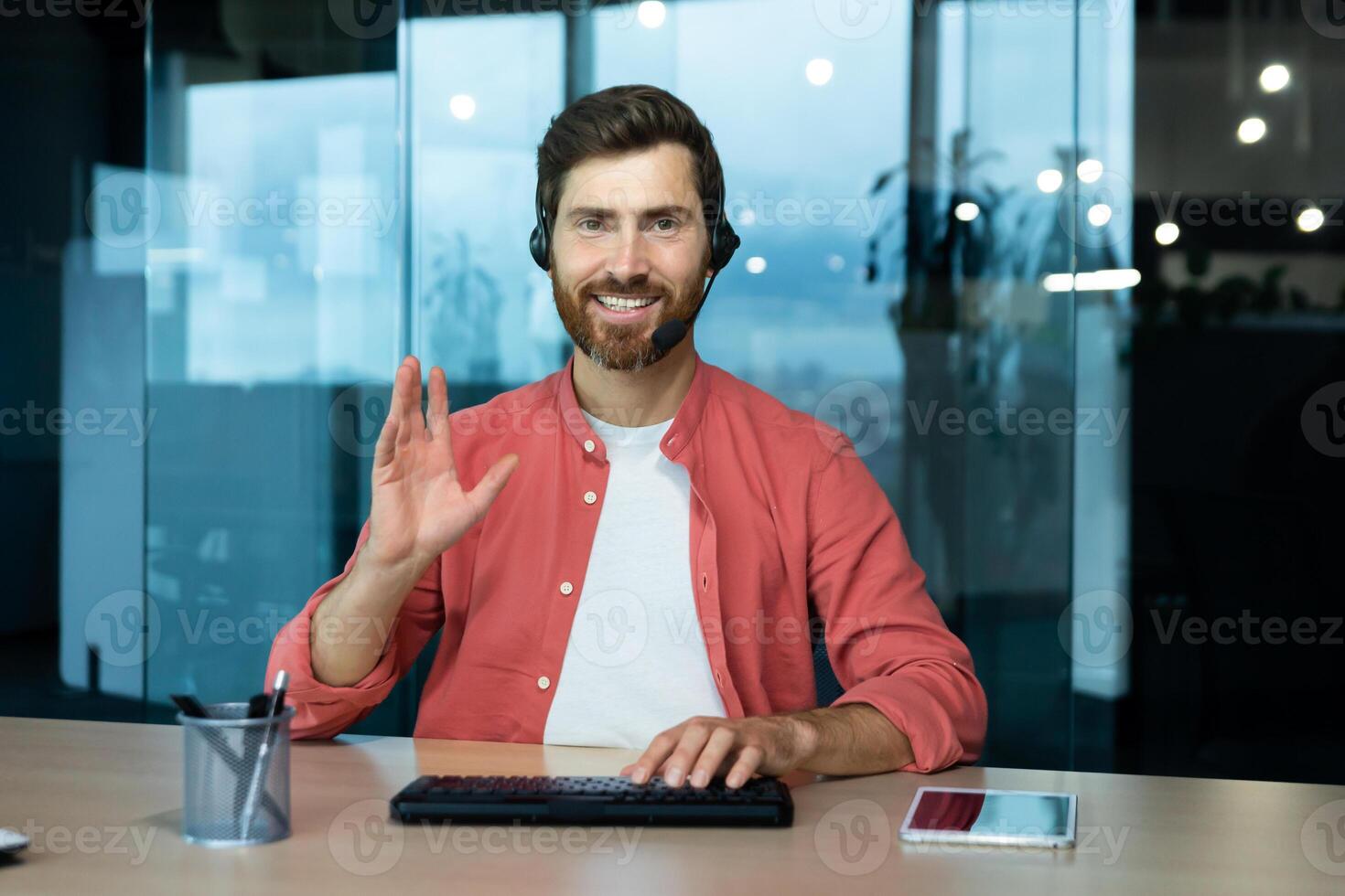 Portrait of a young man in a headset of a psychologist, psychotherapist, sitting in the office at the table with a laptop and consulting online, giving a lecture, looking at the camera, smiling. photo