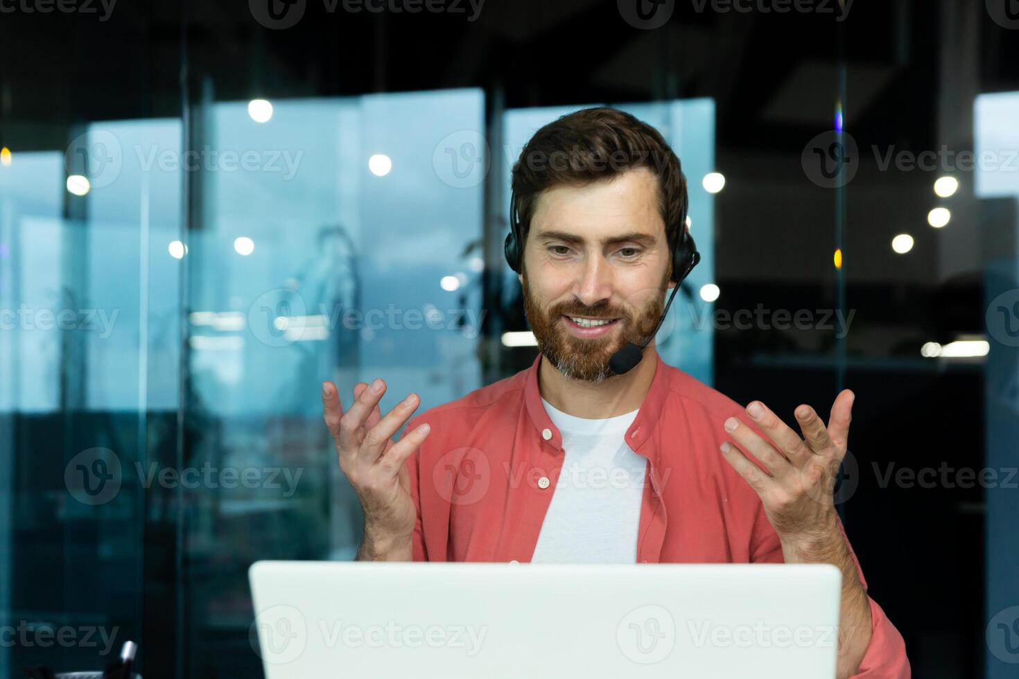 Distance Learning. Portrait of a young man teacher in a headset in a red shirt sits in the office at the table, works on a laptop online, teaches through a call. Explains with his hands, smiles photo