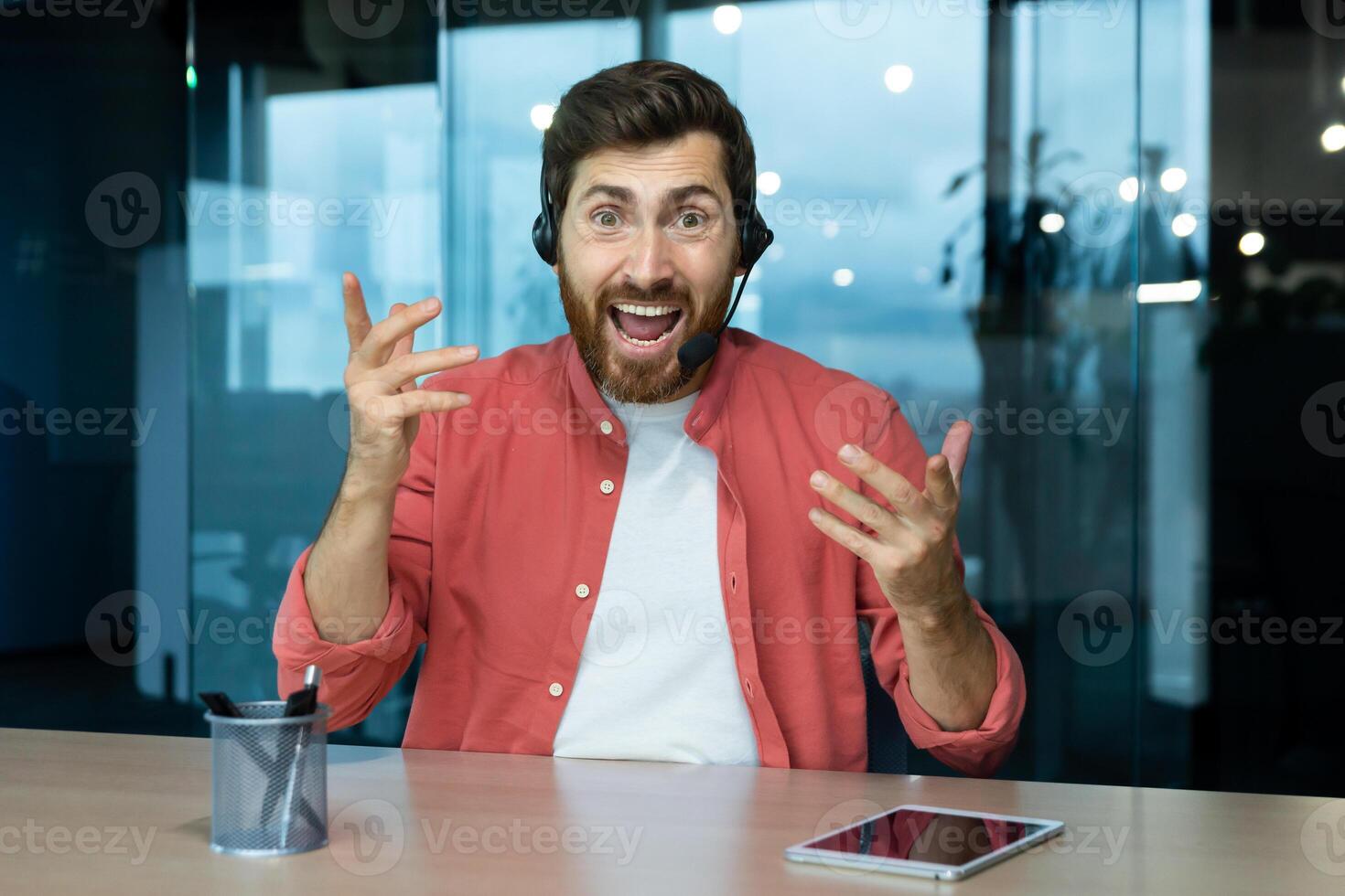 Portrait of a young man in a headset who is excitedly talking to the camera, waving his hands expressively. Sits in the office at the table, communicates via call, consults, commentator. photo