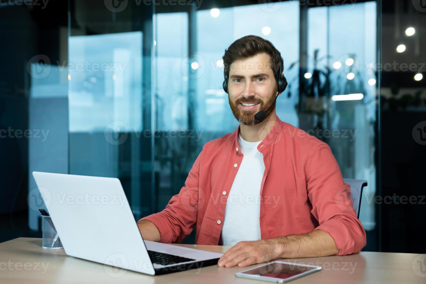 Portrait of mature businessman in red shirt inside office, man with beard and call headset smiling and looking at camera, online customer support worker with laptop. photo