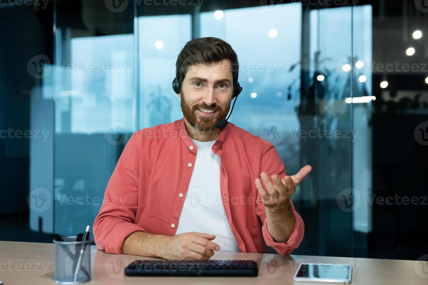 Portrait of a young male blogger in a headset sitting at a desk in the office with a camera and talking via call, recording a podcast, developing his online channel. photo
