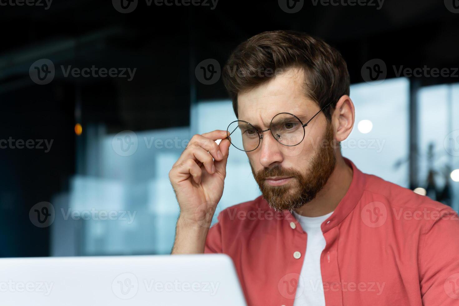 Close-up photo. Portrait of a young male programmer, a businessman who is concentrating on working in the office at a laptop, looking at the monitor, holding glasses on his face. photo