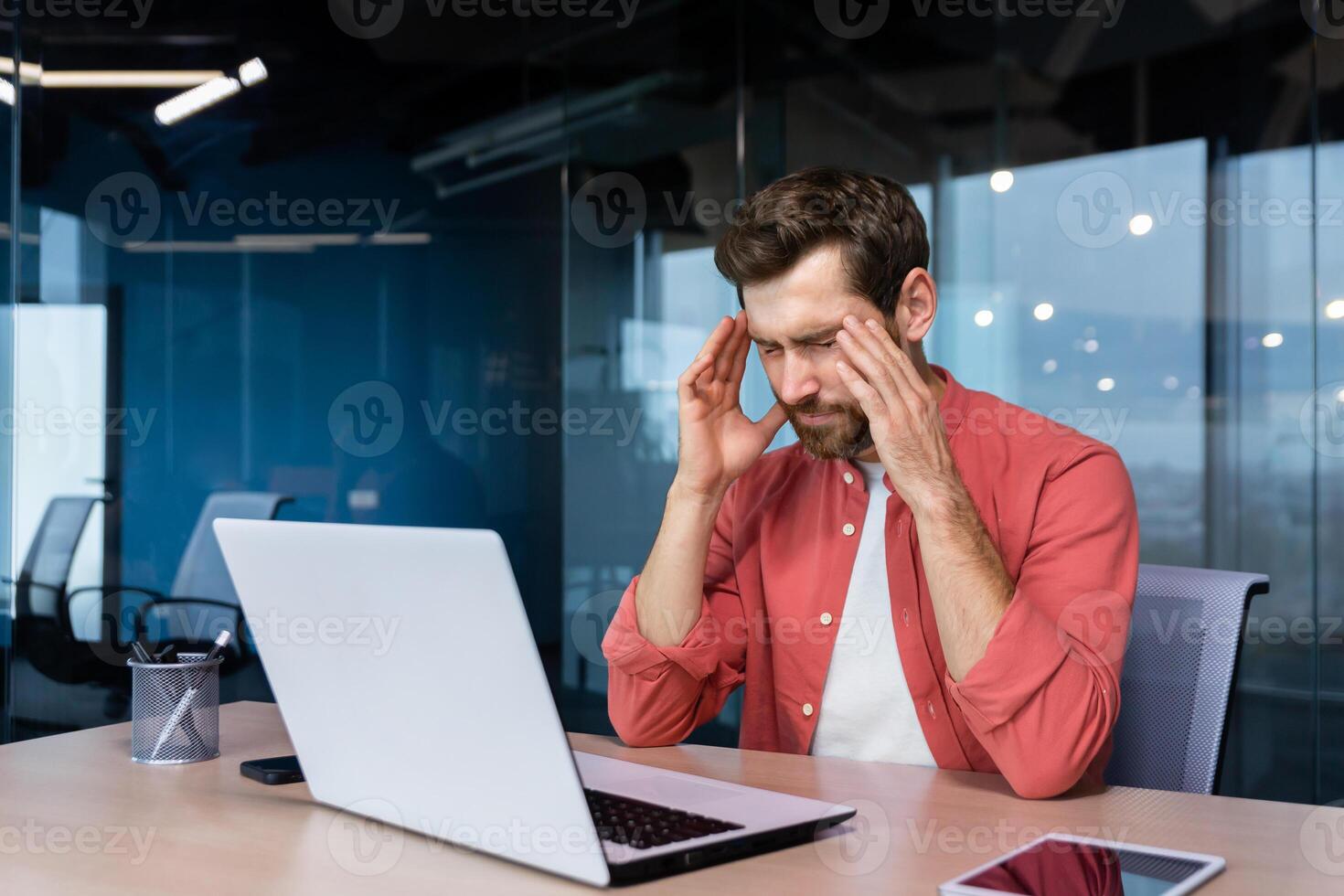 Stress and headache at work. A young businessman in a red shirt is sitting at the desk in the office, holding his head, close eyes. He feels bad, worries about problems, a project, overtime, photo