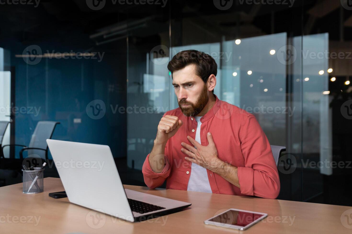 Sick man at workplace, mature worker in red shirt coughing, businessman inside office at work using laptop. photo