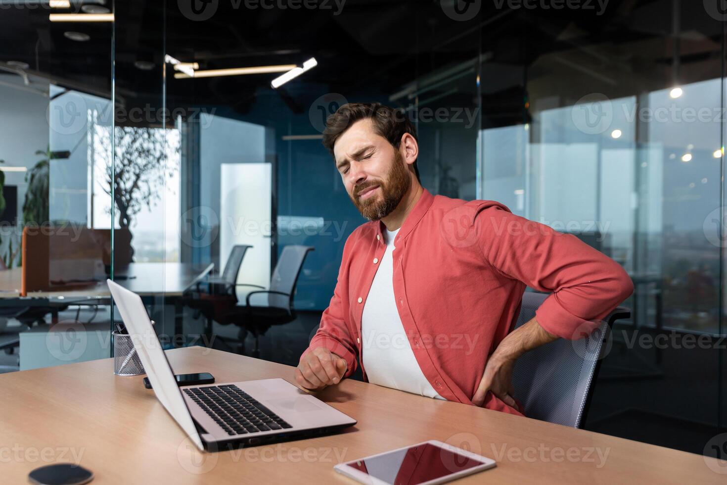 Back pain at the workplace. A young male programmer, freelancer sits in the office at the table and holds his back twisted, stretched, tension pain. Tired from an overtime day. photo