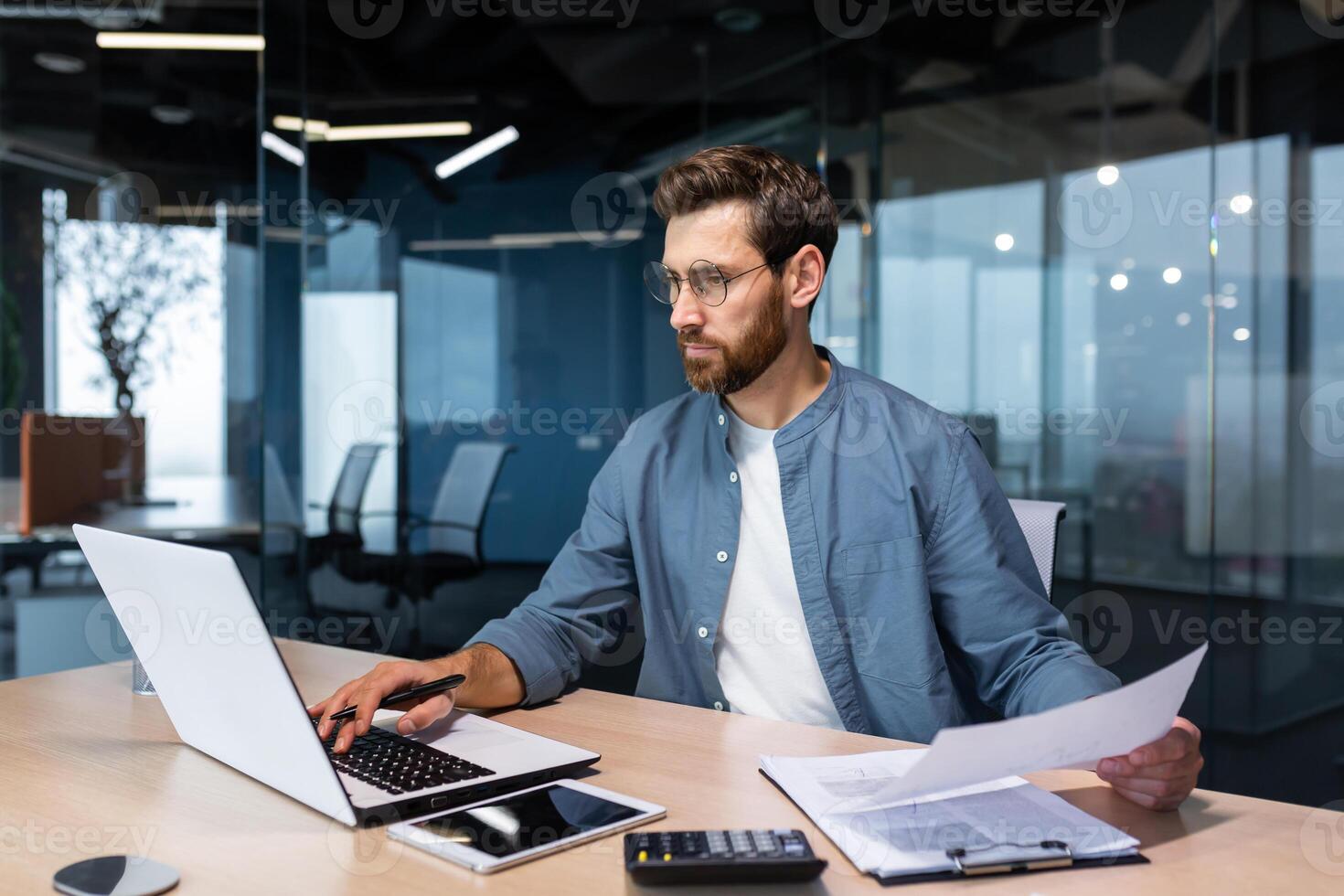 A serious young man accountant, financier, analyst, auditor sits in the office at the table. He holds documents and a pen in his hands, checks accounts, finances, types on a laptop. photo