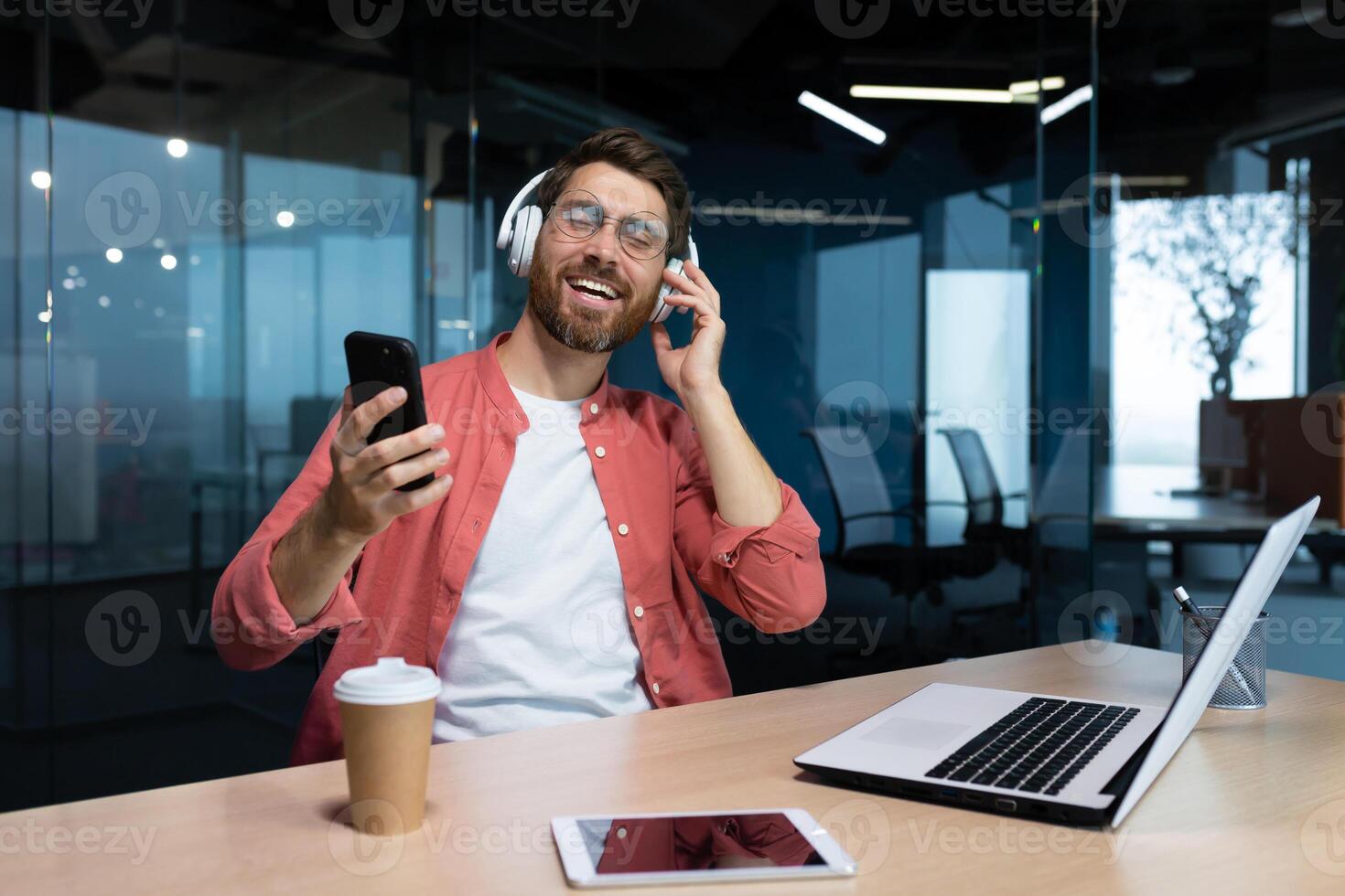 A young male student in headphones studies in the office, campus, at home in front of a laptop, holds a phone in his hand, dances, listens to a lecture. Smiling, resting. photo