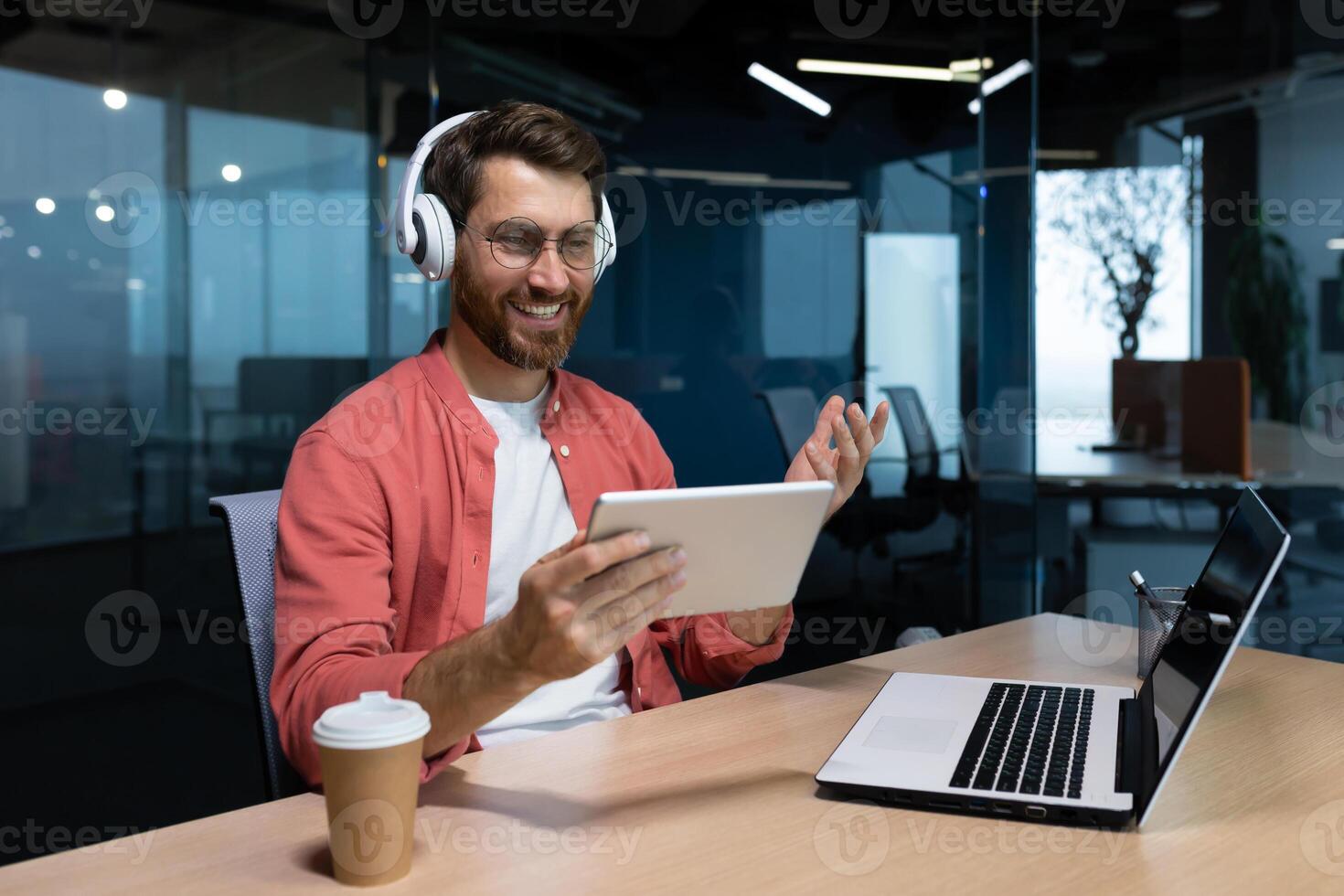 A young businessman freelancer, student, programmer sits in the office at the table with a laptop in headphones, holds and uses a tablet. Talks on a call, listens to music. photo