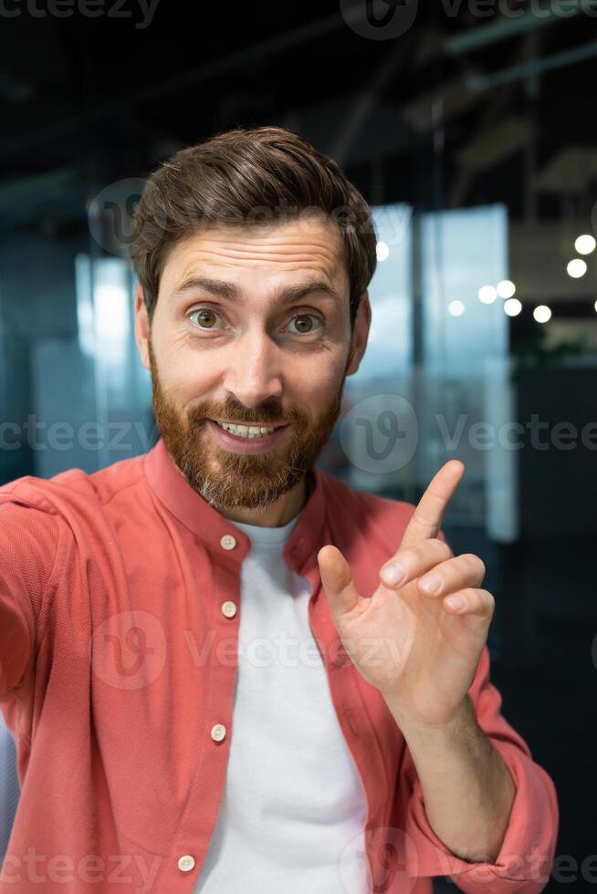 A businessman looks into the camera of the phone, pov. man inside office talking with friends smiling using app on smartphone for call. Vertical shot. photo