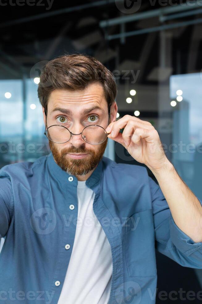 Close-up photo. A young male teacher, trainer, tutor holds a phone in his hands, looks and talks to the camera, puts his hand down and glasses on his face. Vertical photo. photo