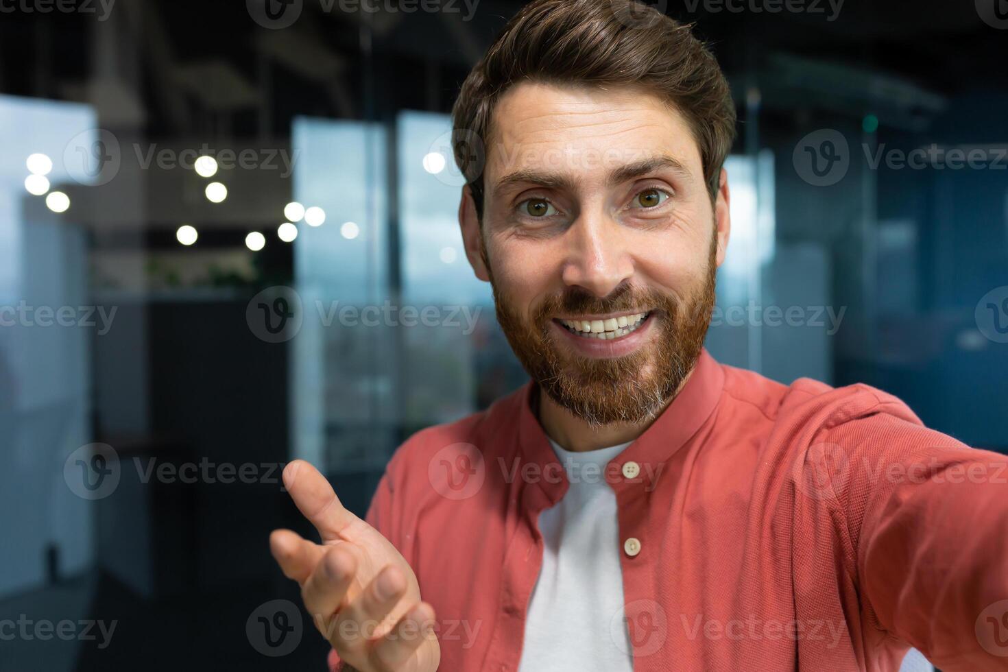 Close-up photo. Young businessman, freelancer, blogger in a red shirt in the office talking on a call from the phone, smiling at the camera. photo