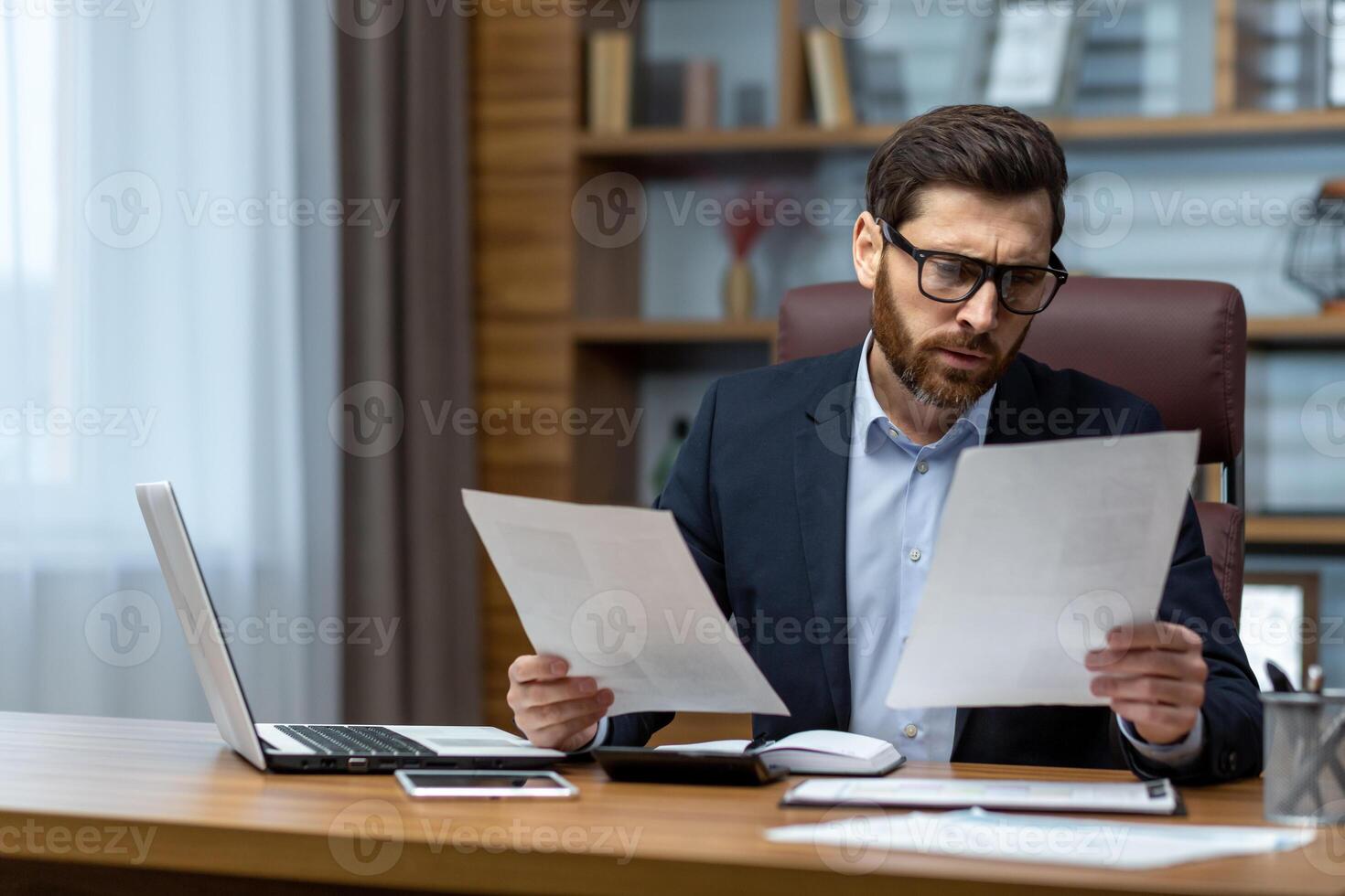 Serious and focused financier accountant on paper work inside office, mature man using calculator and laptop for calculating reports and summarizing accounts, businessman at work . photo