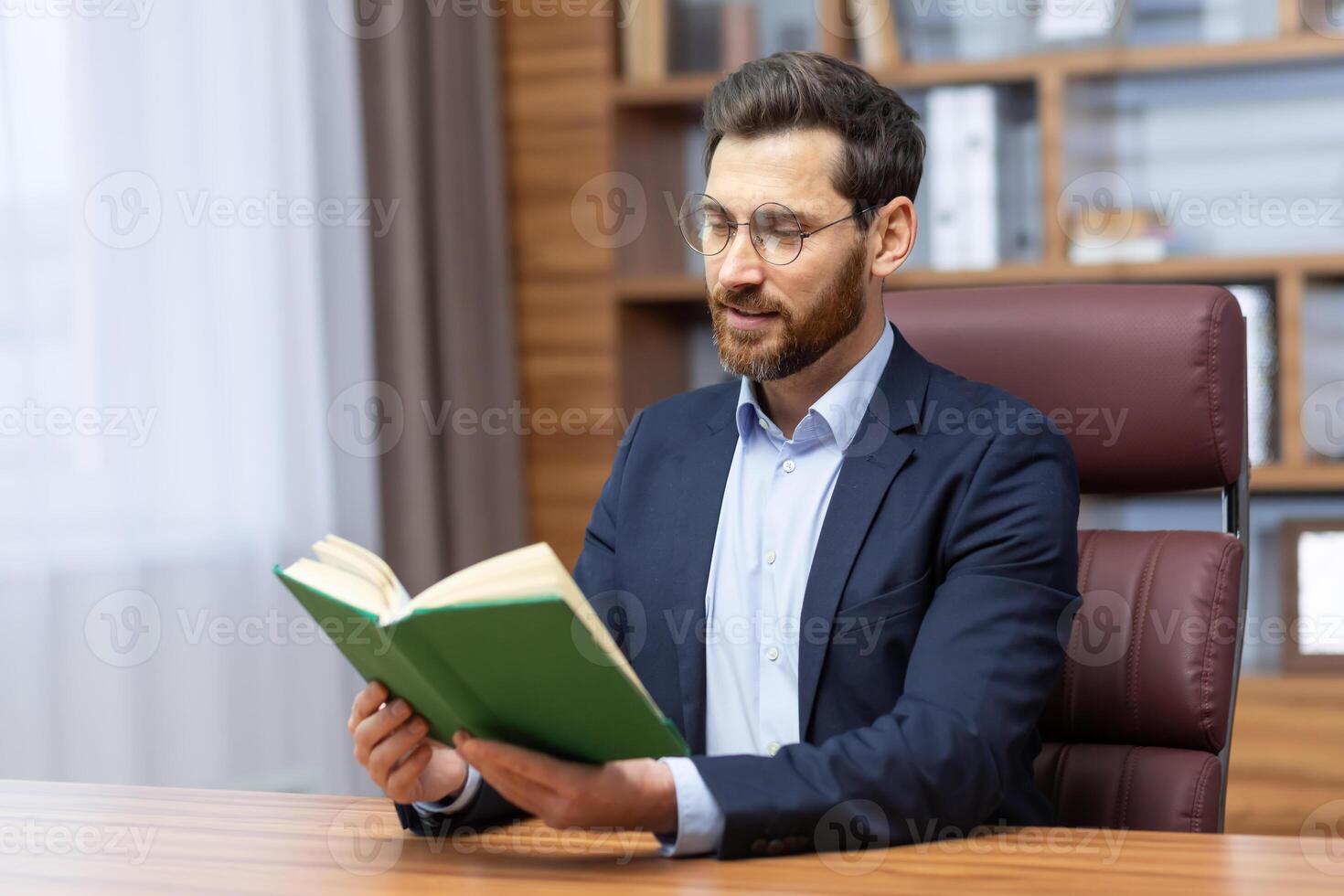 Leisure businessman resting and reading book sitting at desk in business suit inside home office, joyful man improving his financial knowledge. photo