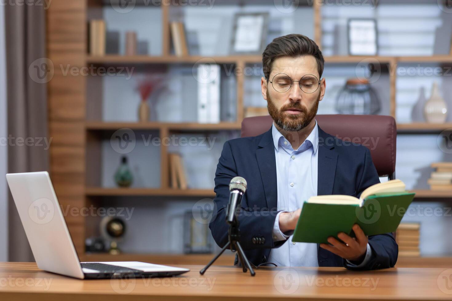 Distance Learning. A young male teacher sits in the office at a table in front of a laptop and a microphone and reads an online lecture from a book to students, pupils. photo
