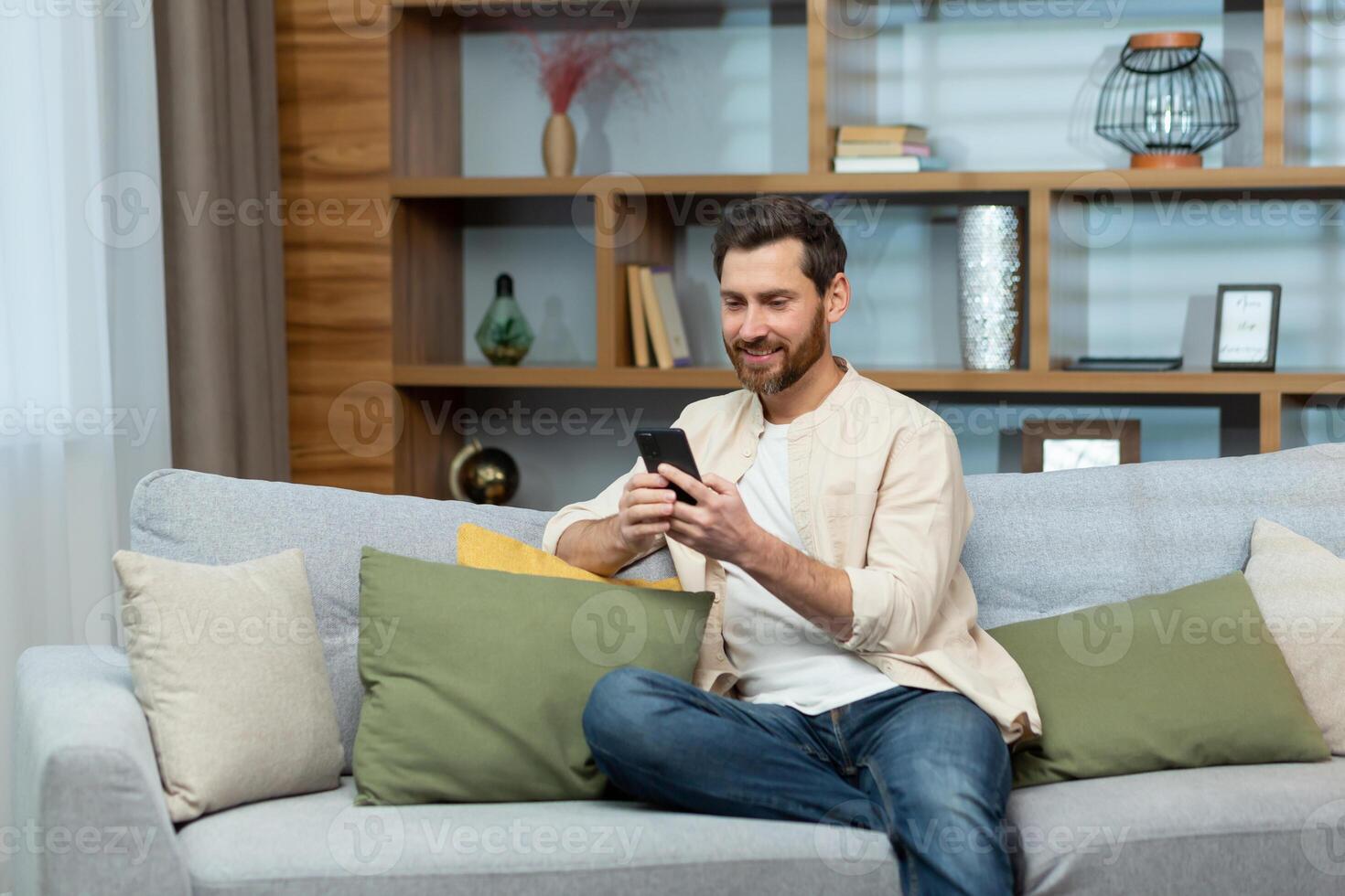 A mature man with a beard in a shirt at home is sitting on the sofa in the living room, using the phone, typing messages and browsing the Internet online. photo