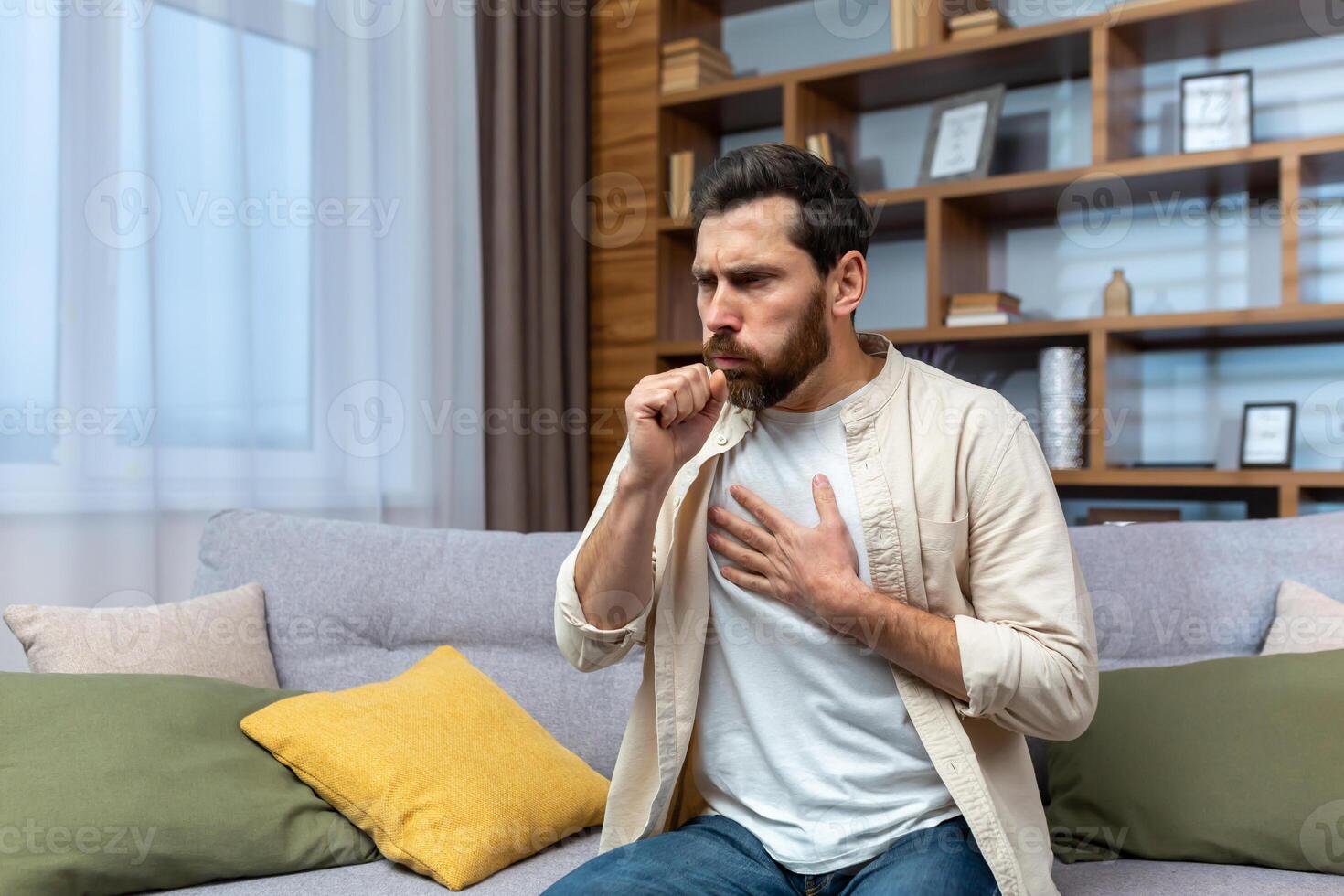 Sick mature man alone at home sitting on sofa coughing holding hands to chest in living room. photo