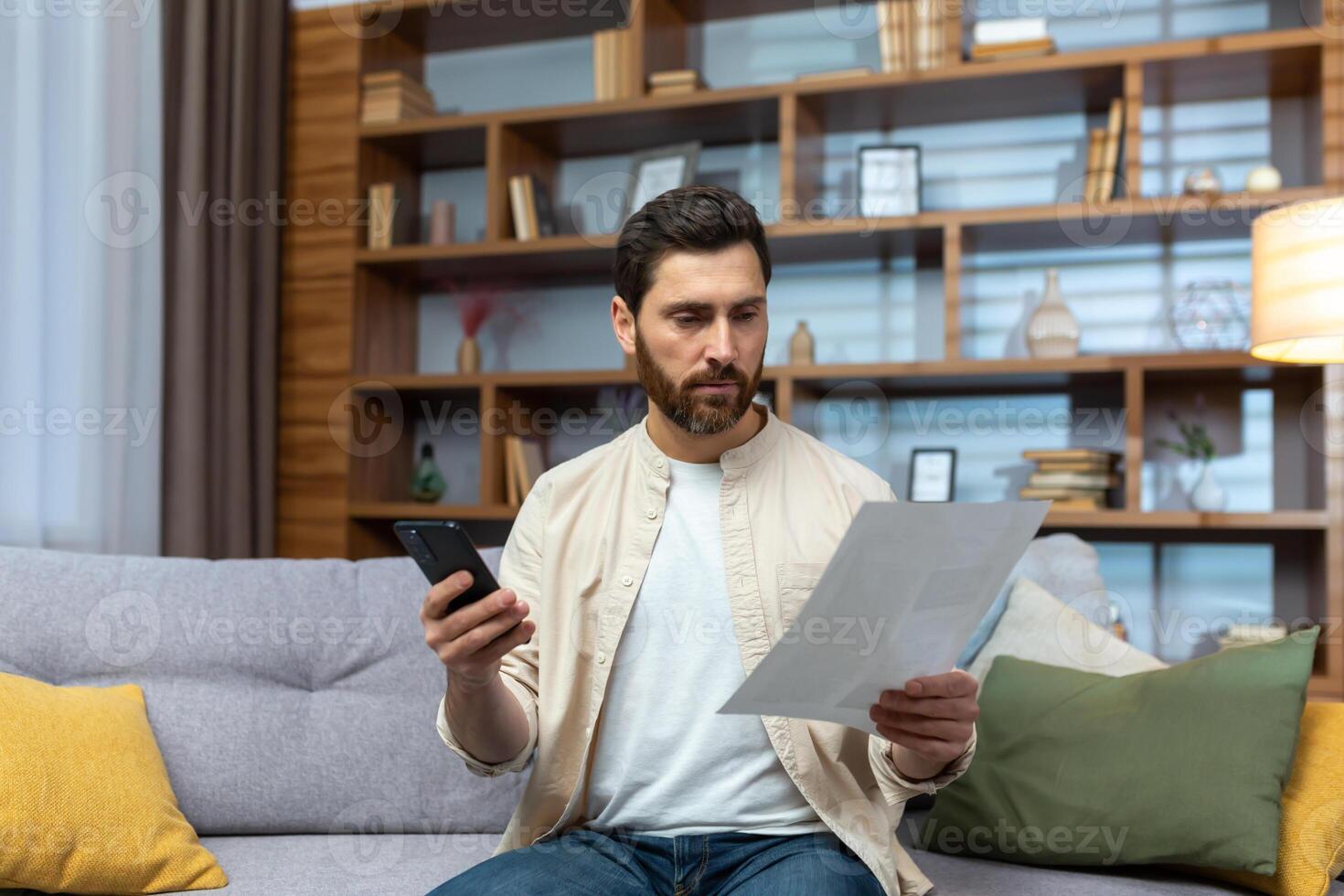 A worried young man is sitting on the sofa at home, holding a phone and documents, a letter. Checks the agreement, accounts, credit. photo