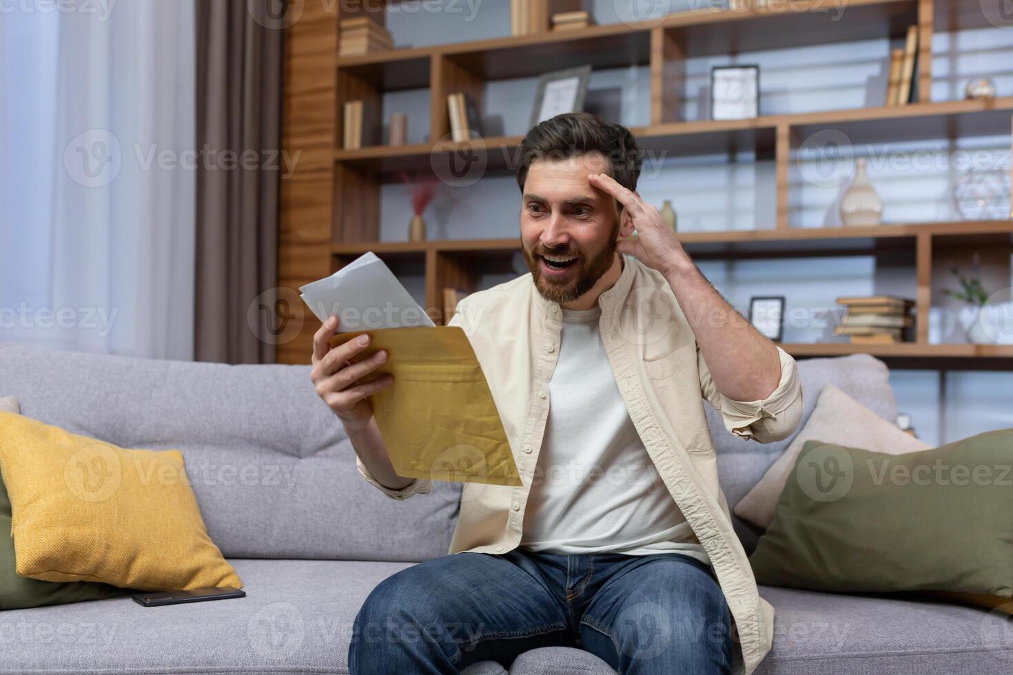 Joyful man at home holding envelope with notification message reading and smiling happy with news sitting on sofa inside living room. photo