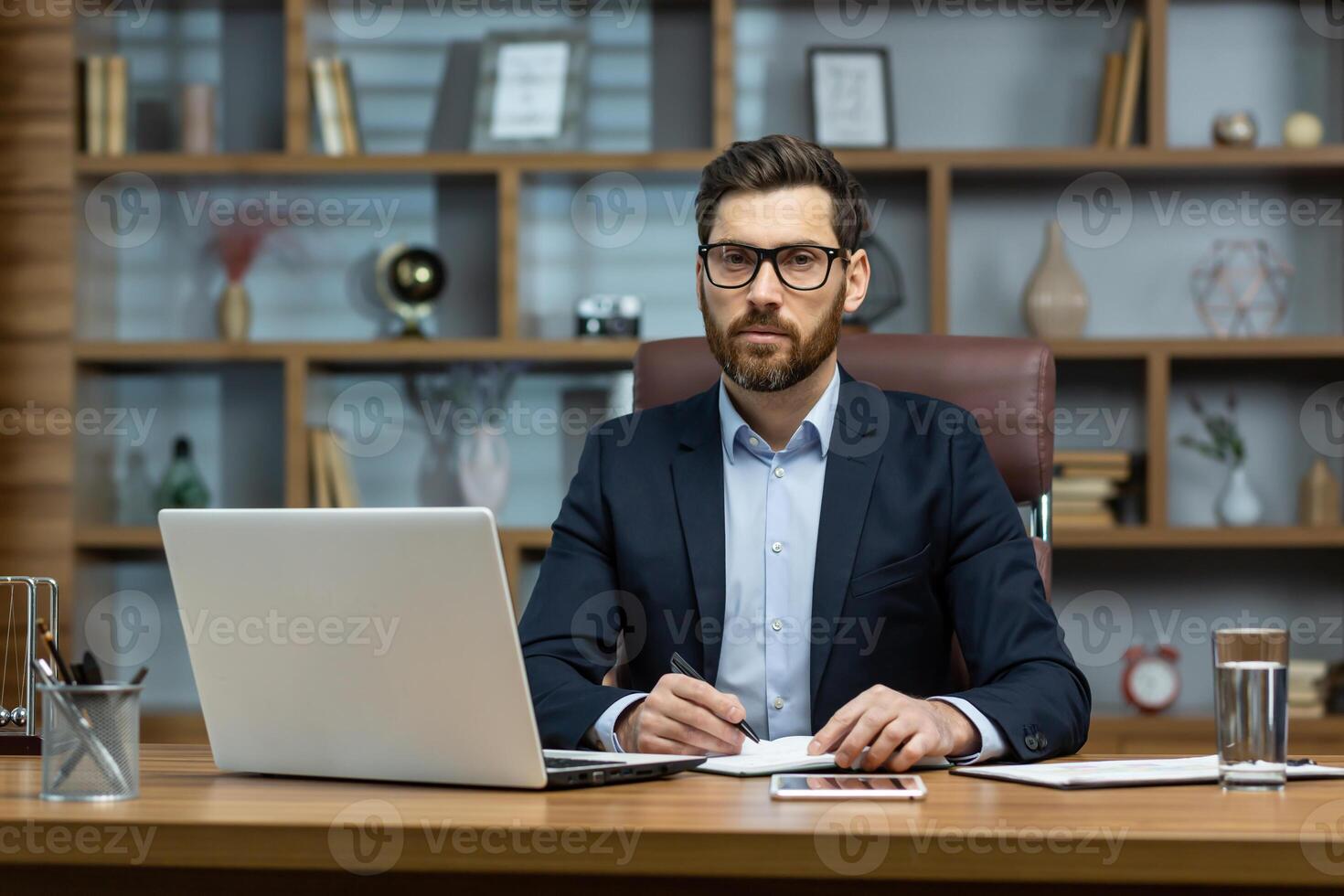 Serious thinking boss inside the office portrait of a mature businessman with a beard and glasses, the man is looking at the camera with concentration, working with documents on a laptop at the table photo