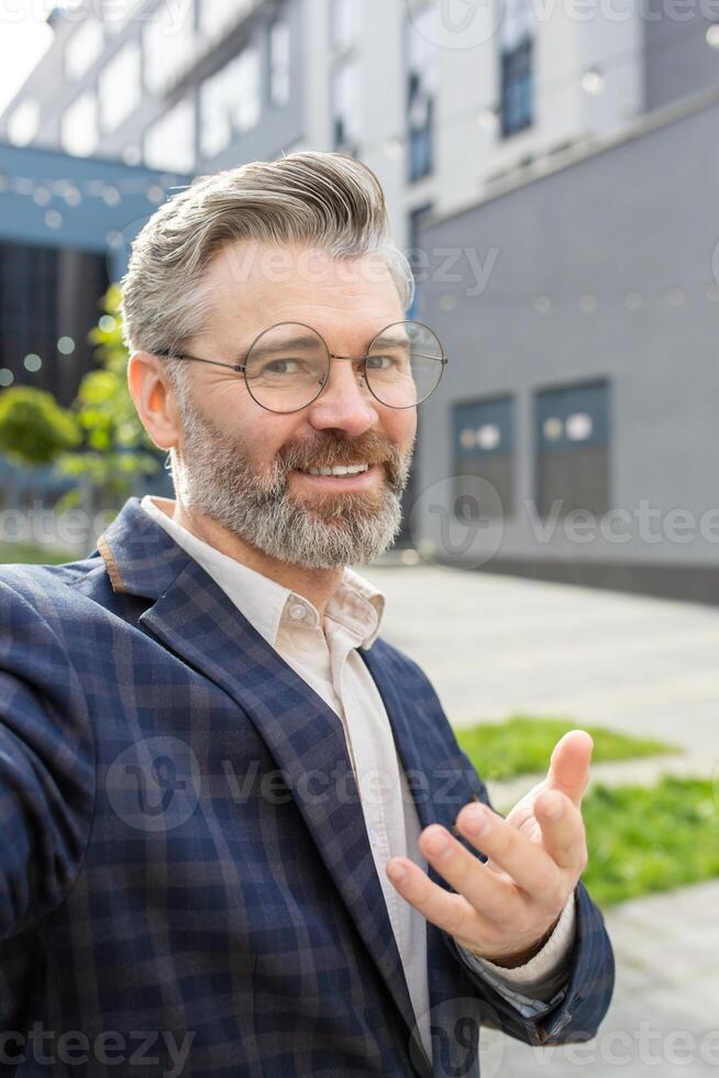 Portrait of a friendly senior businessman with a confident smile, gesturing as if to converse, standing outside an urban office building. photo