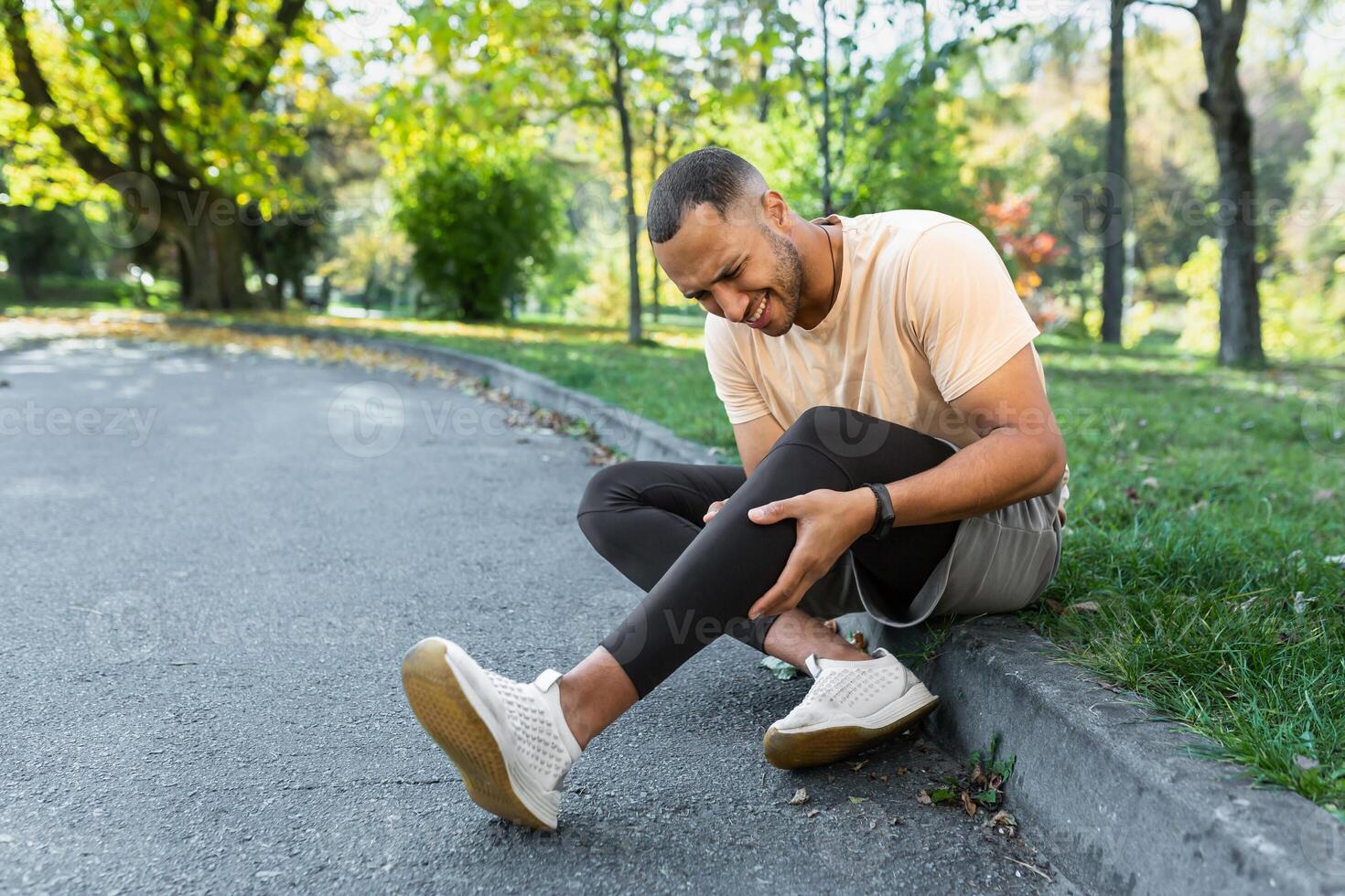 Runner injured his leg while jogging in the park near the trees, hispanic man has muscle spasm massaging his knee, man is running and exercising fitness on a sunny day. photo